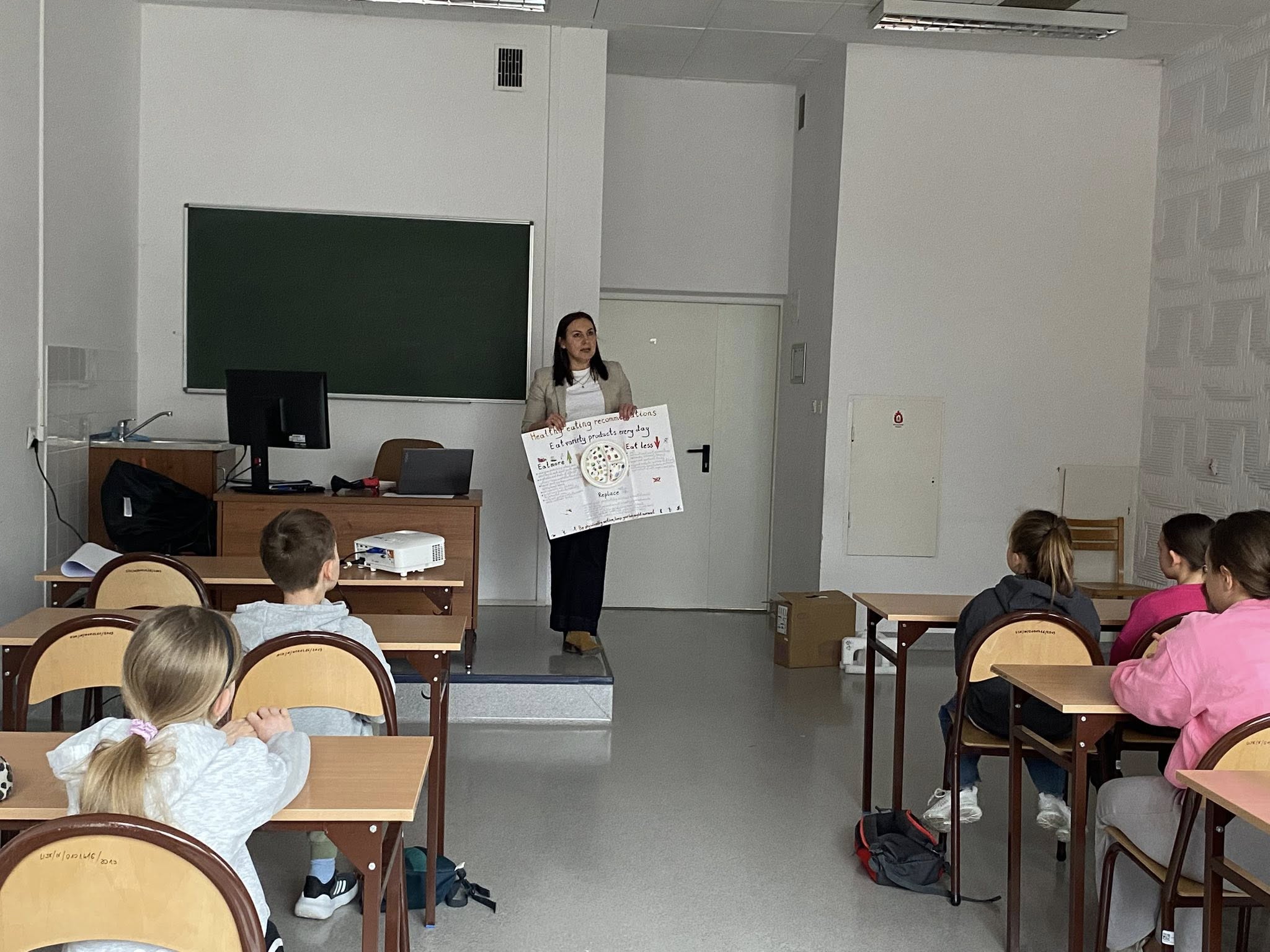 Sala lekcyjna, w której uczestnicy siedzą przy ławkach i obserwują prezentację prowadzoną z przodu. Osoba prowadząca pokazuje plakat z ilustracjami i omawia jego treść przy tablicy. Pomieszczenie ma jasne ściany, oświetlenie sufitowe oraz podstawowe wyposażenie szkolne.