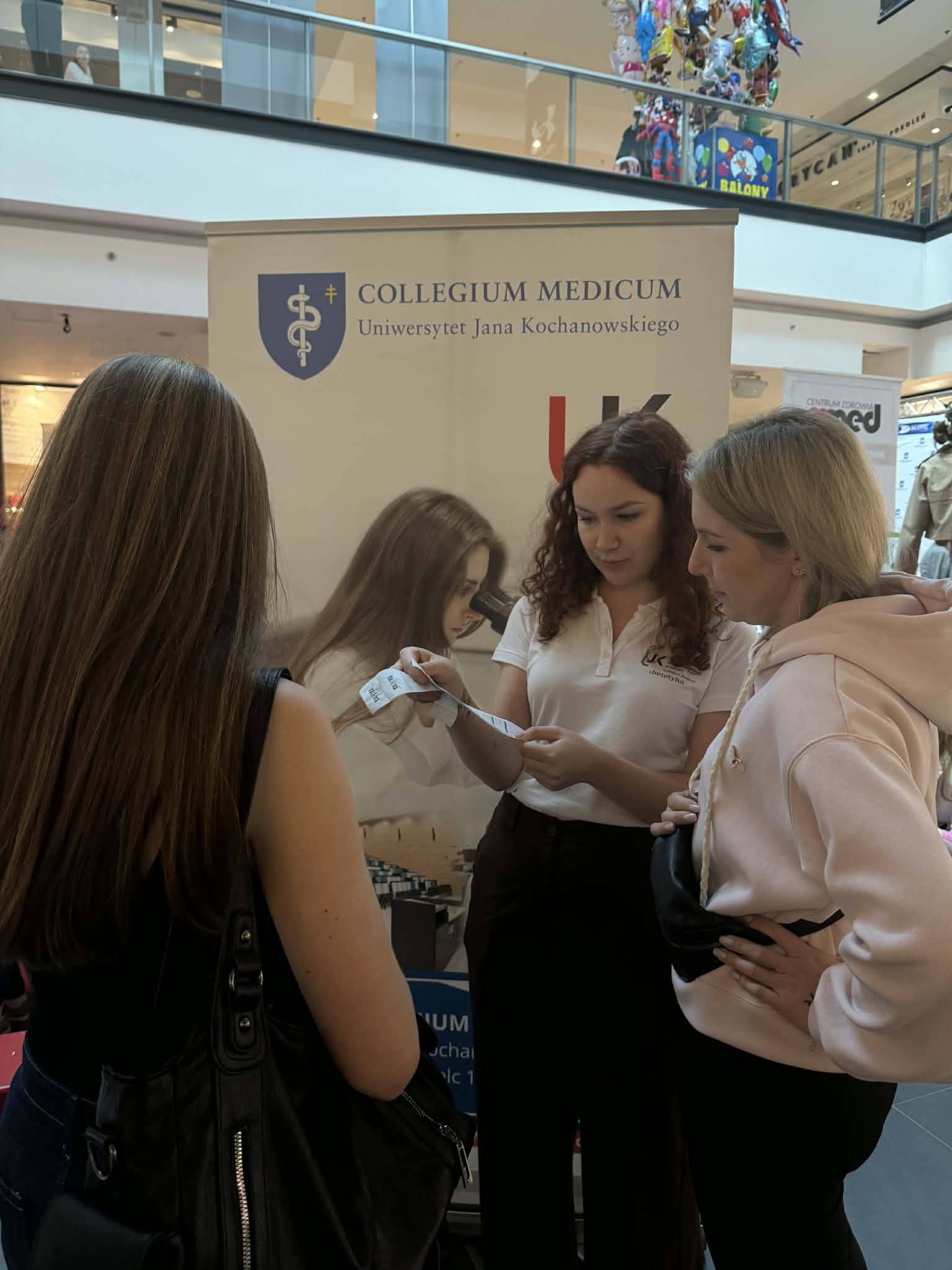 Trzy osoby stoją przy stoisku informacyjnym w przestrzeni publicznej, omawiając materiały promocyjne. W tle widoczne są elementy galerii handlowej oraz inne osoby, co wskazuje na wydarzenie informacyjne lub akcję promocyjną.