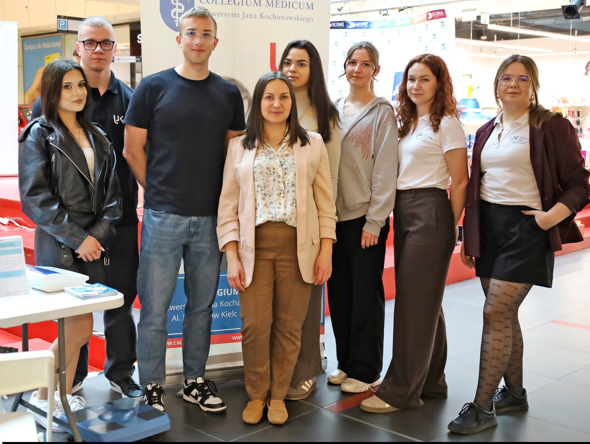 Grupa uczestników stoi w pomieszczeniu przed banerem promocyjnym, pozując do wspólnego zdjęcia. W tle widoczne są elementy wydarzenia, takie jak materiały informacyjne na stole, co wskazuje na spotkanie lub akcję promocyjną w przestrzeni publicznej.