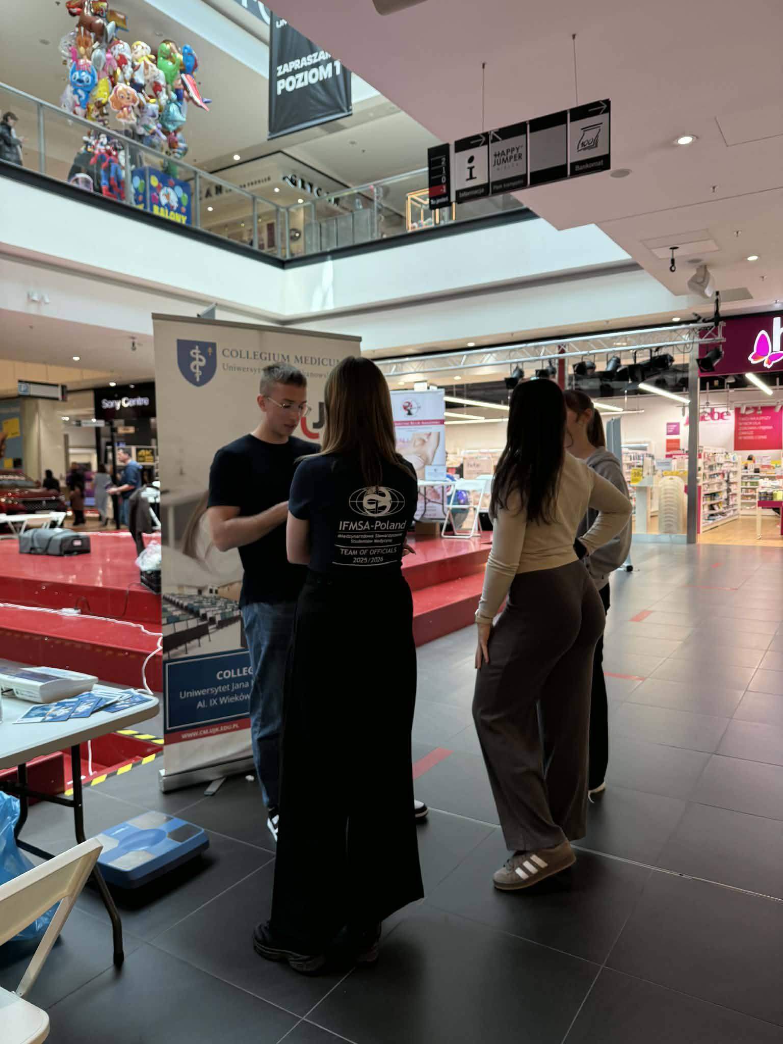 Grupa uczestników rozmawia przy stoisku promocyjnym w galerii handlowej, gdzie prezentowane są materiały informacyjne uczelni i organizacji. W tle widoczne są sklepy oraz elementy przestrzeni handlowej, co wskazuje na wydarzenie informacyjne lub akcję promocyjną.