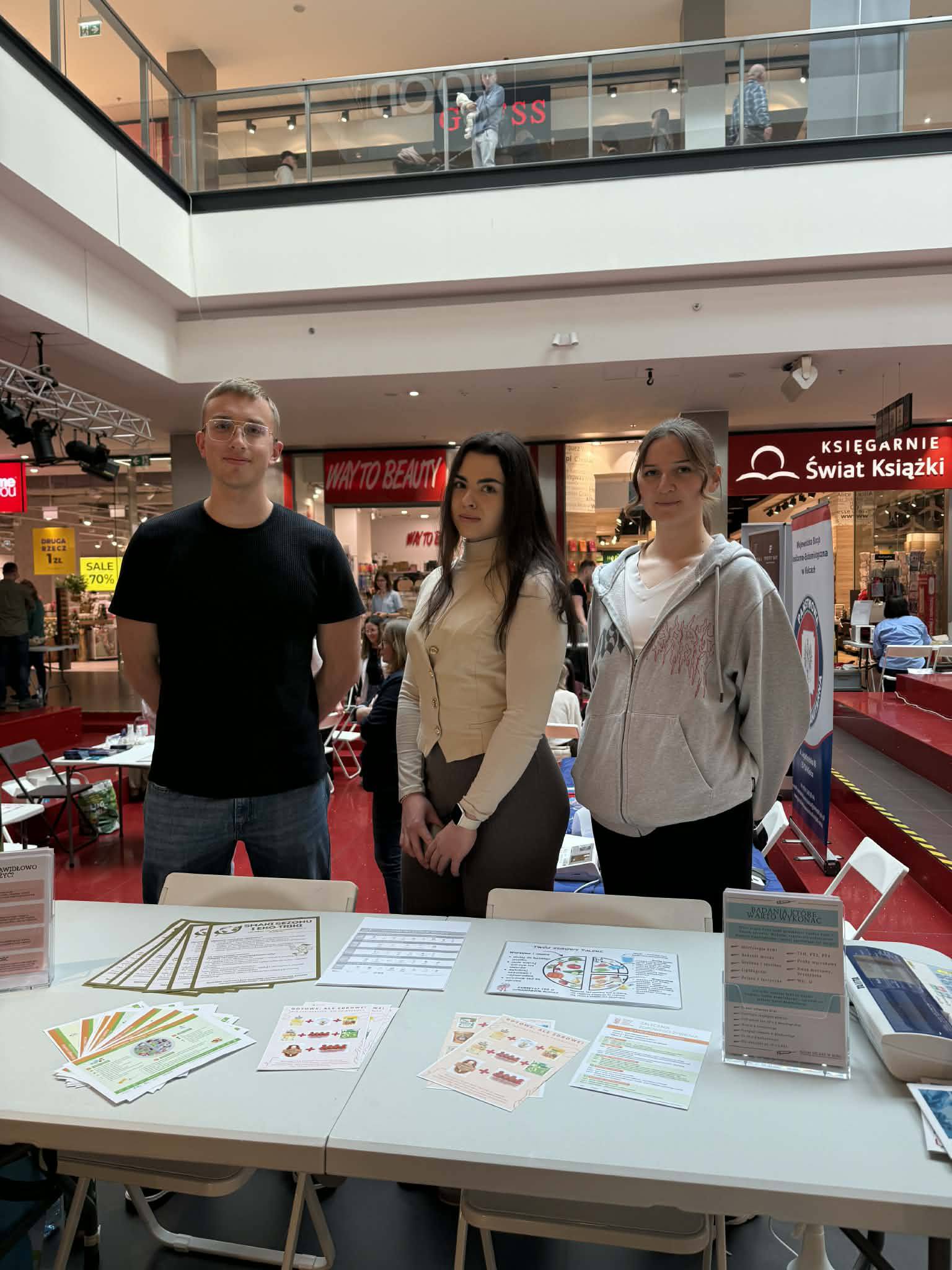 Trzy osoby stoją za stołem z materiałami informacyjnymi, prowadząc stoisko w galerii handlowej. Na blacie znajdują się ulotki i kolorowe materiały promocyjne. W tle widoczne są sklepy i oznaczenia lokali, co wskazuje na wydarzenie promocyjne w przestrzeni handlowej.