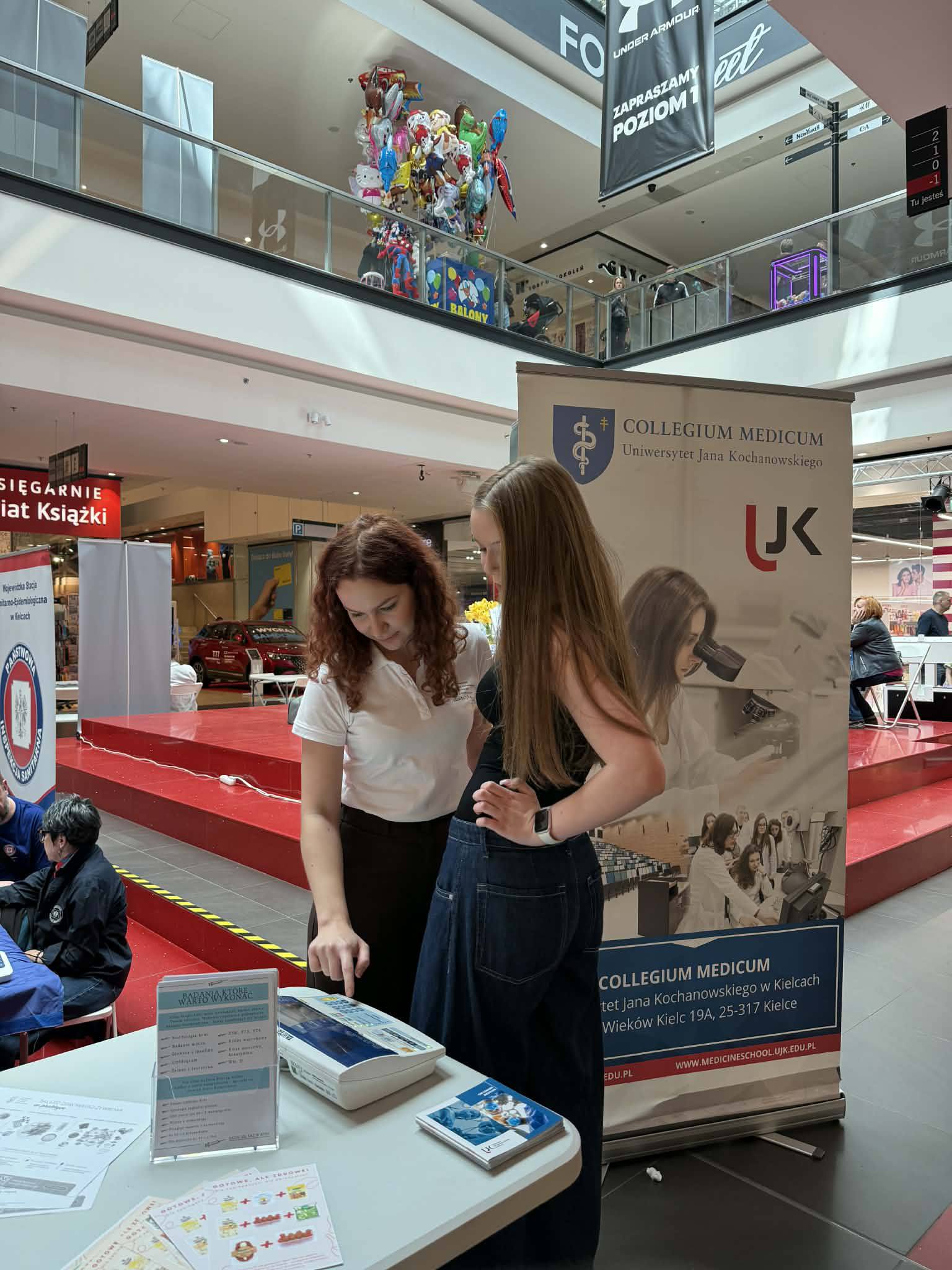 Dwie osoby stoją przy stoisku informacyjnym w przestrzeni handlowej, omawiając materiały znajdujące się na stole z ulotkami. W tle widoczny jest baner uczelni oraz elementy galerii handlowej, takie jak balustrady, schody i inne stoiska, co wskazuje na wydarzenie promocyjne.