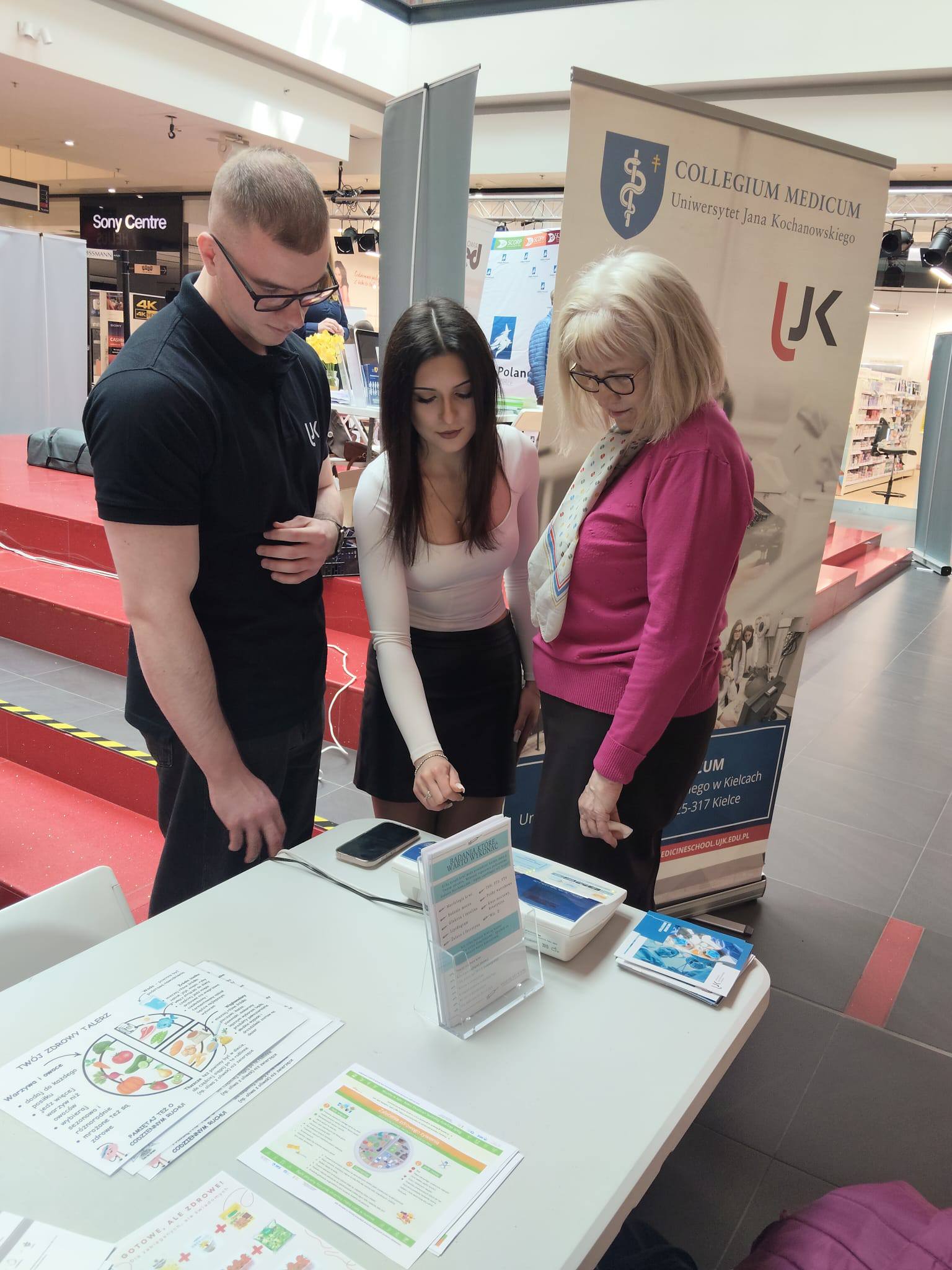 Trzy osoby stoją przy stoisku informacyjnym w galerii handlowej i omawiają materiały promocyjne rozłożone na stole. W tle widoczny jest baner uczelni, a jasna przestrzeń z przeszklonym dachem wskazuje na wydarzenie informacyjne lub promocyjne.