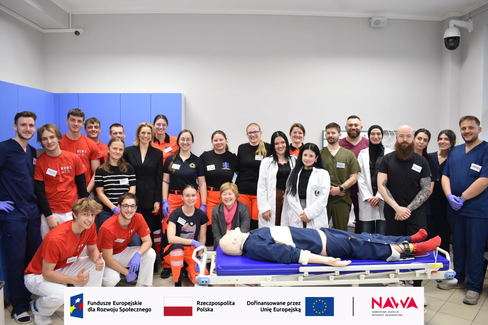 Grupa uczestników stoi w sali szkoleniowej o charakterze medycznym, pozując do zdjęcia wokół łóżka z fantomem wykorzystywanym do ćwiczeń. Wnętrze wyposażone jest w sprzęt i szafki medyczne, co wskazuje na zajęcia praktyczne lub szkolenie.