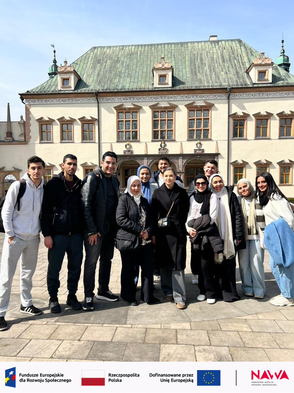 Grupa uczestników stoi przed zabytkowym budynkiem o ozdobnej architekturze, pozując do wspólnego zdjęcia. W tle widoczna jest jasna elewacja i charakterystyczny dach, a słoneczna pogoda podkreśla plenerowy charakter ujęcia.