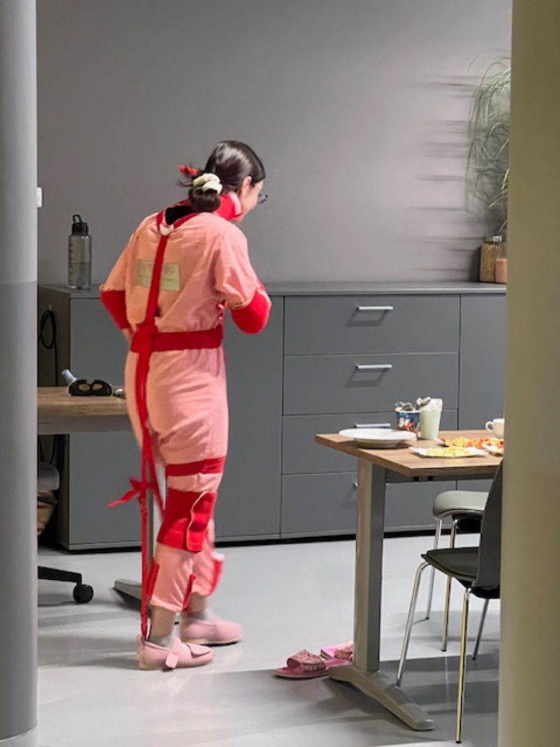 Osoba w specjalnym stroju symulacyjnym stoi w nowoczesnym pomieszczeniu, prezentując ograniczenia ruchowe. W tle widoczna jest zabudowa meblowa z rośliną oraz stół z przygotowanymi naczyniami, co wskazuje na kontekst szkoleniowy lub edukacyjny.