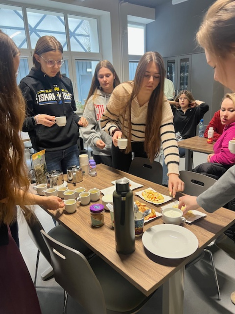 Grupa uczestników gromadzi się wokół stołu z poczęstunkiem, przygotowując napoje i przekąski lub częstując się nimi. Na stole znajdują się kubki, termos, owoce i inne produkty. W tle widoczne są osoby siedzące oraz duże okna wpuszczające światło dzienne, co wskazuje na swobodną atmosferę spotkania.