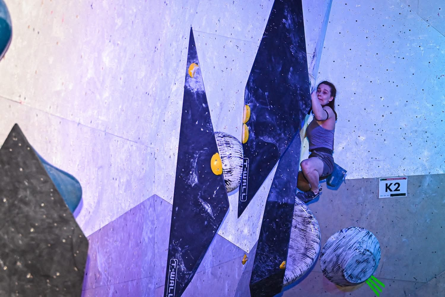 Osoba wspina się po ściance boulderowej, pokonując trasę z dużymi i mniejszymi chwytami. Wykonuje ruchy wymagające siły i precyzji, opierając się o ścianę. Całość odbywa się w hali wspinaczkowej oświetlonej kolorowym światłem, co podkreśla sportowy charakter wydarzenia.