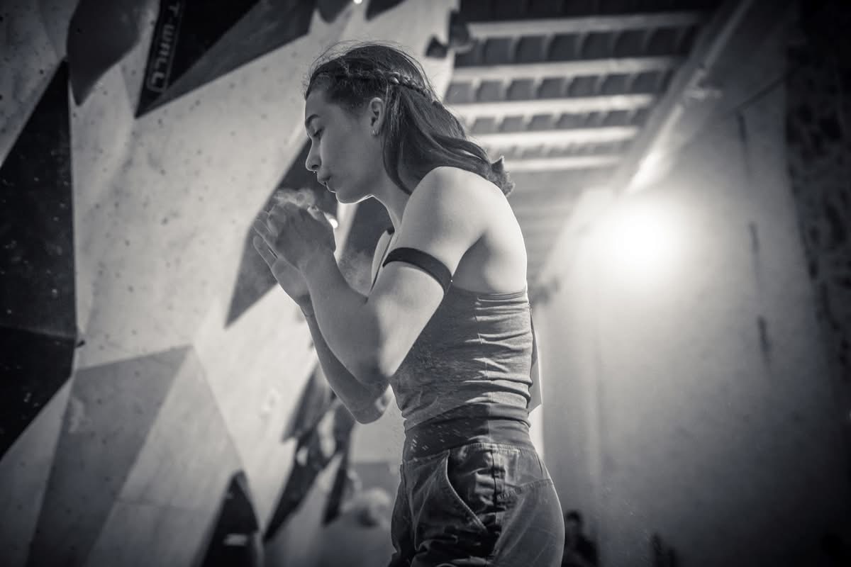 Czarno-białe zdjęcie przedstawiające osobę w hali wspinaczkowej przygotowującą dłonie do wspinaczki, z widocznym pyłem z magnezji unoszącym się w powietrzu. W tle znajduje się fragment ścianki wspinaczkowej, a kontrastowe oświetlenie nadaje scenie dynamiczny charakter.