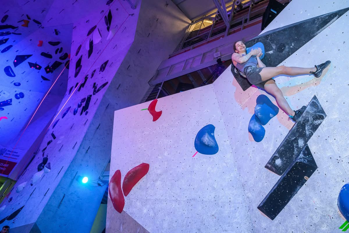 Osoba wspina się po ściance wspinaczkowej z kolorowymi chwytami, pokonując stromy fragment trasy. W tle widoczne są inne sekcje ściany oświetlone kolorowym światłem, co podkreśla dynamiczny i sportowy charakter miejsca.