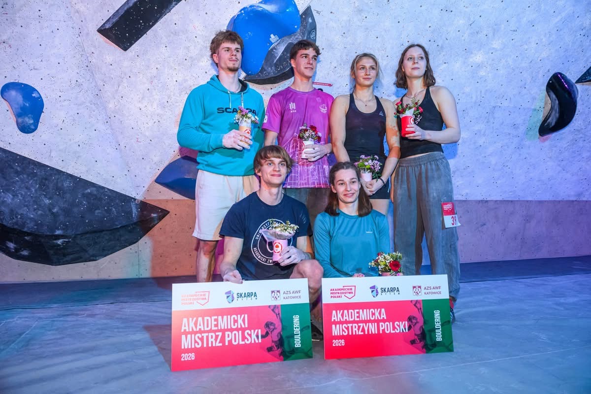 Grupa uczestników pozuje do zdjęcia przed ścianką wspinaczkową z kolorowymi uchwytami. Część osób stoi, a część klęczy z przodu, trzymając drobne upominki, co sugeruje uroczysty lub podsumowujący charakter spotkania.