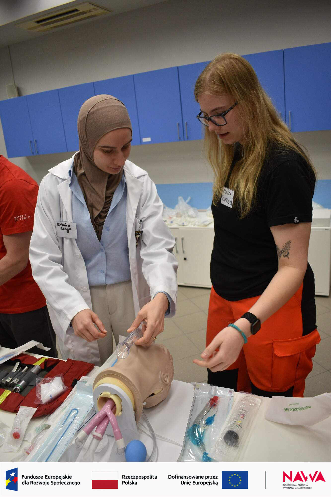 Zajęcia szkoleniowe w sali medycznej, podczas których uczestnicy ćwiczą procedury na fantomie. Na stole znajdują się narzędzia i materiały do nauki, a jedna z osób wykonuje ćwiczenie, podczas gdy druga obserwuje. W tle widoczne są szafki z wyposażeniem.