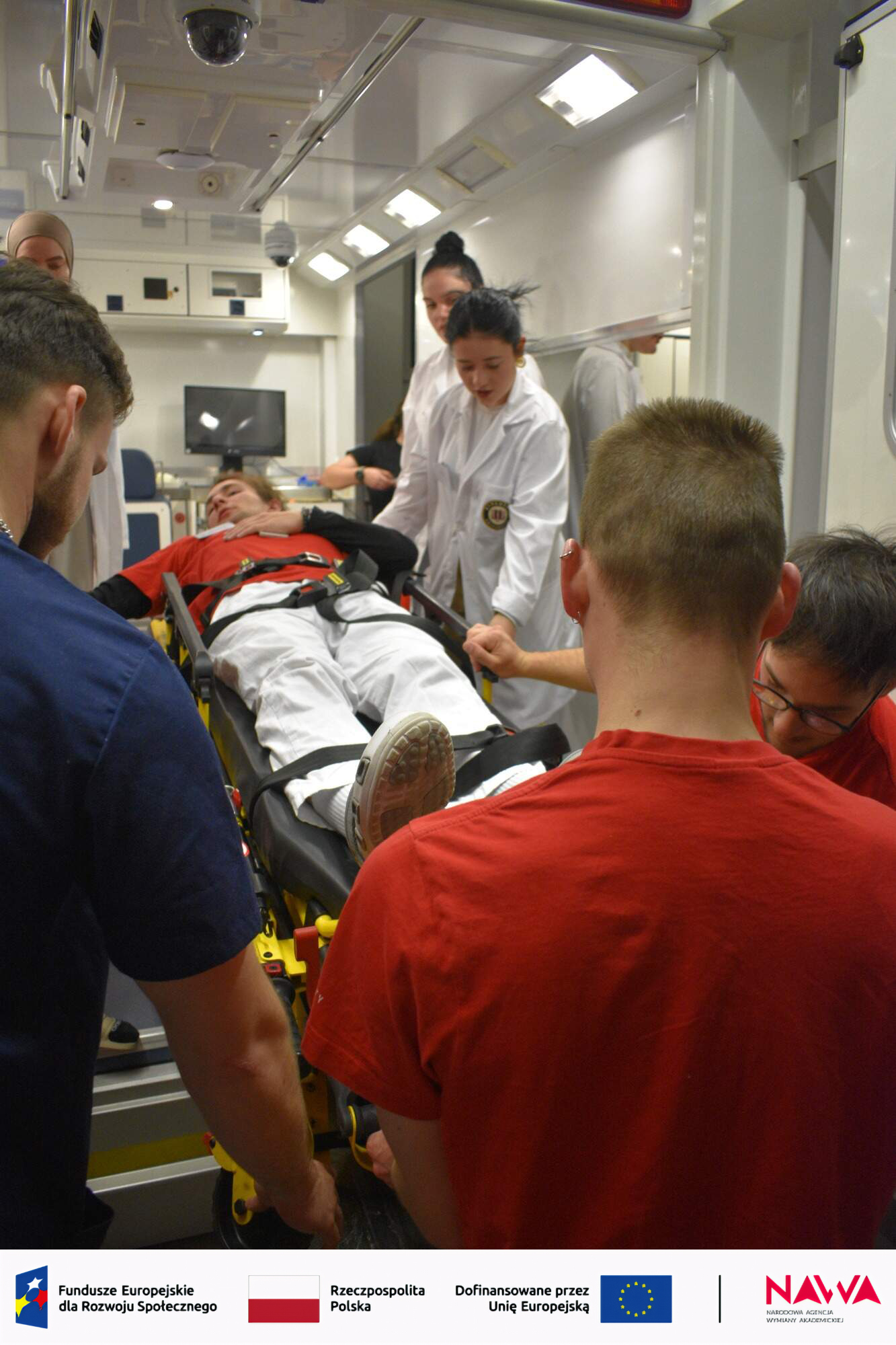 Uczestnicy biorą udział w szkoleniu medycznym w przestrzeni przypominającej ambulans lub salę symulacyjną, skupiając się wokół pacjenta na noszach. Osoba prowadząca omawia przebieg działań, a pozostali obserwują lub uczestniczą w ćwiczeniu. Wnętrze wyposażone jest w sprzęt medyczny i oświetlenie typowe dla środowiska ratunkowego.