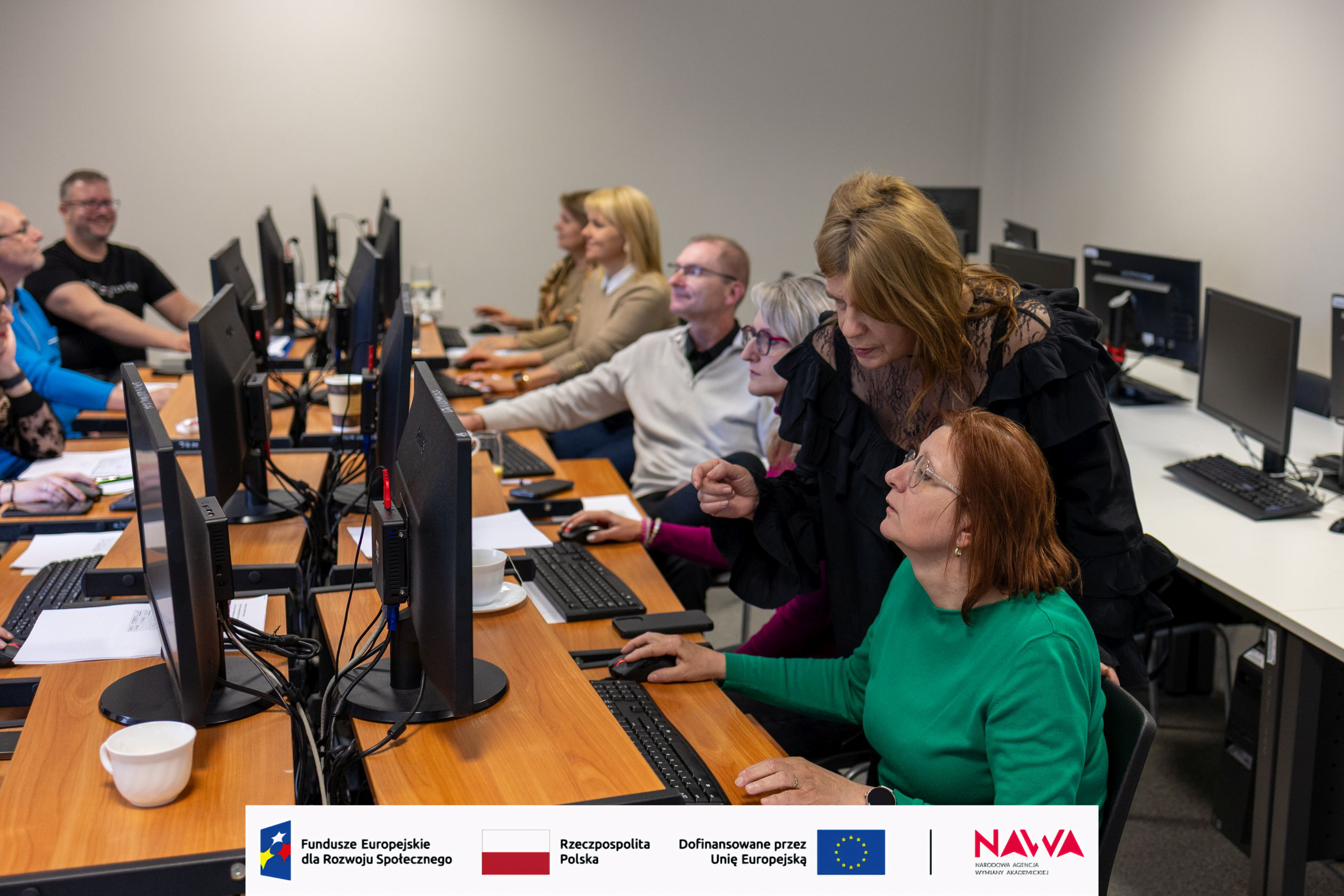 Sala szkoleniowa z długimi biurkami i stanowiskami komputerowymi, przy których uczestnicy pracują i skupiają się na zadaniach. Osoba prowadząca pomaga przy jednym ze stanowisk, wskazując elementy na ekranie. Na blatach znajdują się kubki i materiały, a wnętrze ma neutralny, profesjonalny charakter.