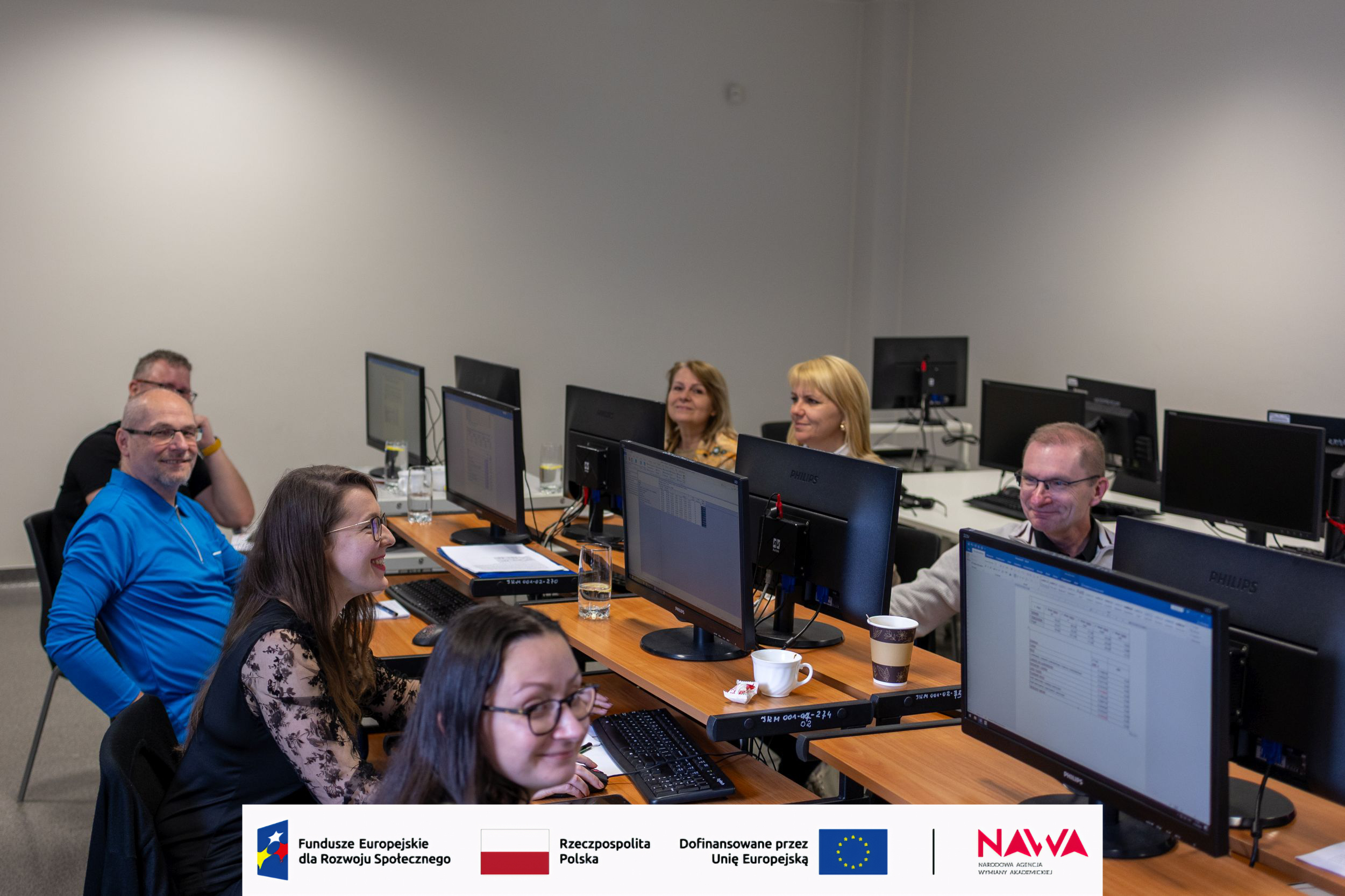 Uczestnicy siedzą przy stanowiskach komputerowych w sali szkoleniowej i pracują na wyświetlanych dokumentach. Na biurkach znajdują się drobne przedmioty, takie jak kubki i notatki. Pomieszczenie jest jasne, uporządkowane i przystosowane do zajęć z wykorzystaniem komputerów.