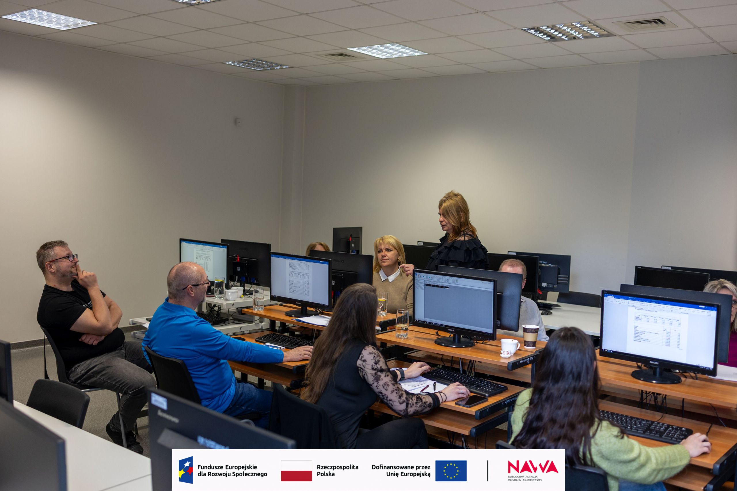 Sala szkoleniowa z komputerami, w której uczestnicy siedzą przy stanowiskach i biorą udział w zajęciach. Osoba prowadząca stoi z przodu i omawia materiał, a pozostali pracują przy monitorach. Wnętrze ma jasne ściany, oświetlenie sufitowe oraz wyposażenie typowe dla pracowni komputerowej.