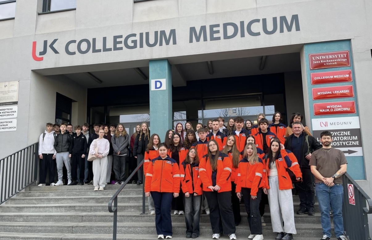 Grupa uczestników stoi na schodach przed budynkiem Collegium Medicum, pozując do wspólnego zdjęcia. W tle widoczne jest wejście do budynku z oznaczeniami oraz tablicami informacyjnymi, co wskazuje na uczelniany charakter miejsca.