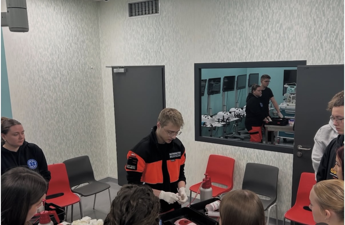 Uczestnicy biorą udział w szkoleniu z zakresu ratownictwa medycznego, obserwując demonstrację procedury przy stole z materiałami i sprzętem. Osoba prowadząca wykonuje czynności w rękawicach, a pozostali śledzą przebieg zajęć. Wnętrze ma jasne ściany, drzwi oraz lustro odbijające dodatkową przestrzeń z wyposażeniem i stanowiskami komputerowymi.