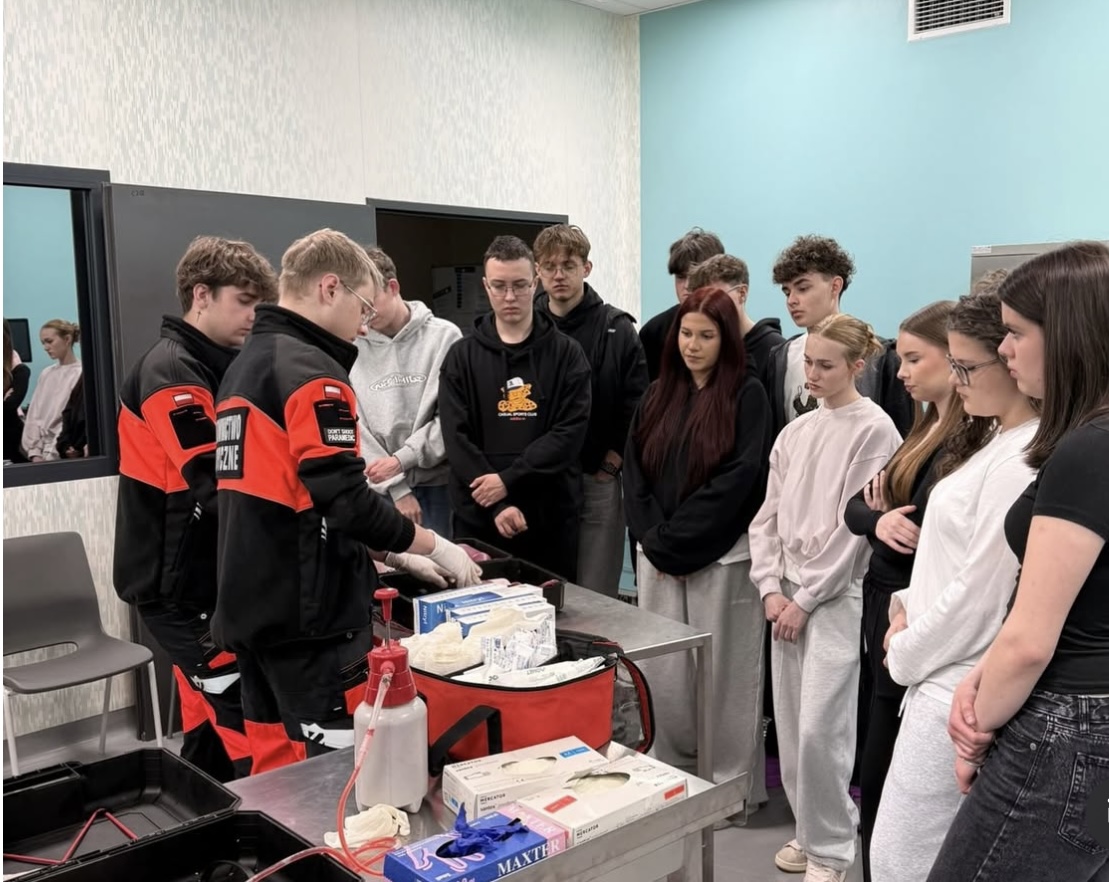 Uczestnicy zgromadzeni w sali szkoleniowej obserwują pokaz. Na stole znajdują się materiały i sprzęt do pierwszej pomocy, wykorzystywane podczas demonstracji. Wnętrze ma jasne ściany i okno, co wskazuje na edukacyjny charakter zajęć.