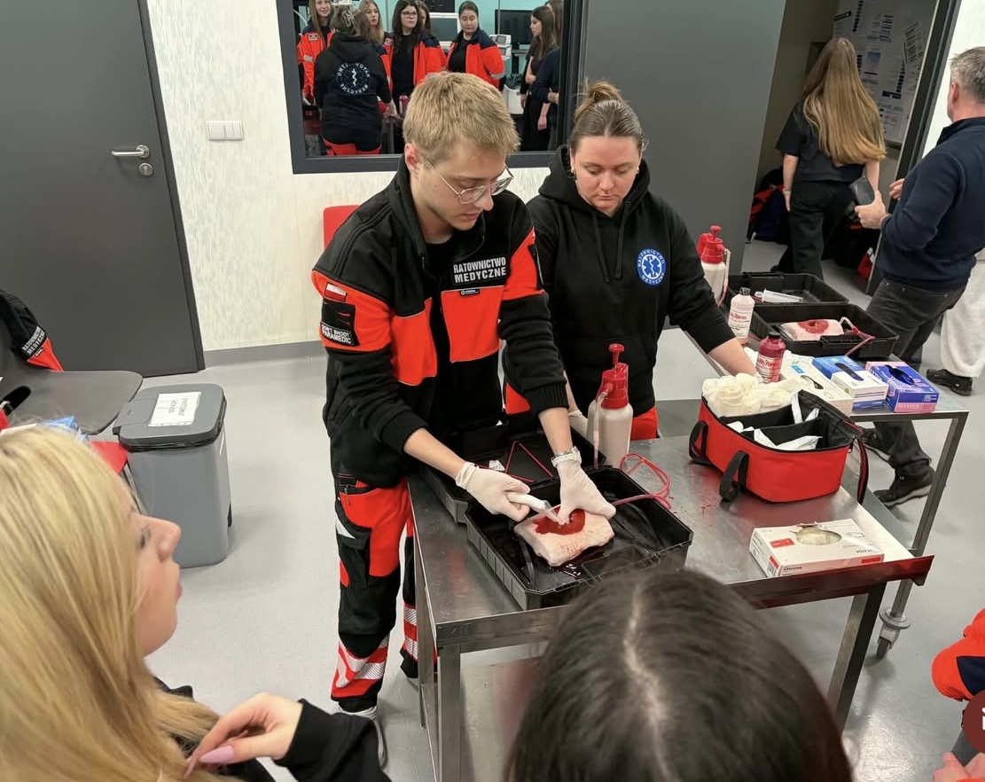 Uczestnicy biorą udział w szkoleniu medycznym, wykonując ćwiczenia na materiale symulującym tkanki. Na stole znajdują się zestawy ratunkowe z opatrunkami i sprzętem, a osoby prowadzące pracują w rękawicach. W tle widoczne jest lustro odbijające pozostałych uczestników oraz wyposażenie sali, co podkreśla praktyczny charakter zajęć.