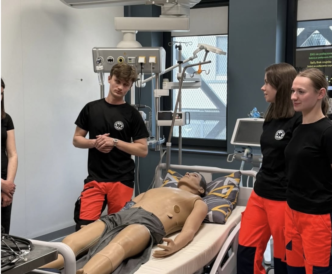 Uczestnicy biorą udział w szkoleniu medycznym, pracując przy łóżku z fantomem pacjenta wyposażonym w czujniki. W sali znajdują się urządzenia monitorujące oraz wyposażenie typowe dla środowiska klinicznego. Pomieszczenie jest dobrze oświetlone, z dużymi oknami i elementami infrastruktury szkoleniowej.