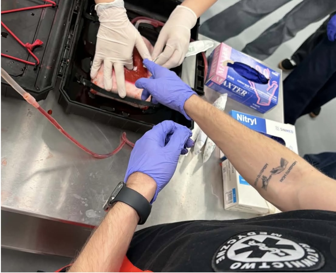 Zbliżenie na ćwiczenia medyczne z wykorzystaniem modelu narządu wypełnionego płynem symulującym krew. Osoby prowadzą i wykonują procedurę w rękawicach, korzystając z zestawu szkoleniowego. Obok widoczne są opakowania rękawic oraz sprzęt wykorzystywany podczas zajęć praktycznych.