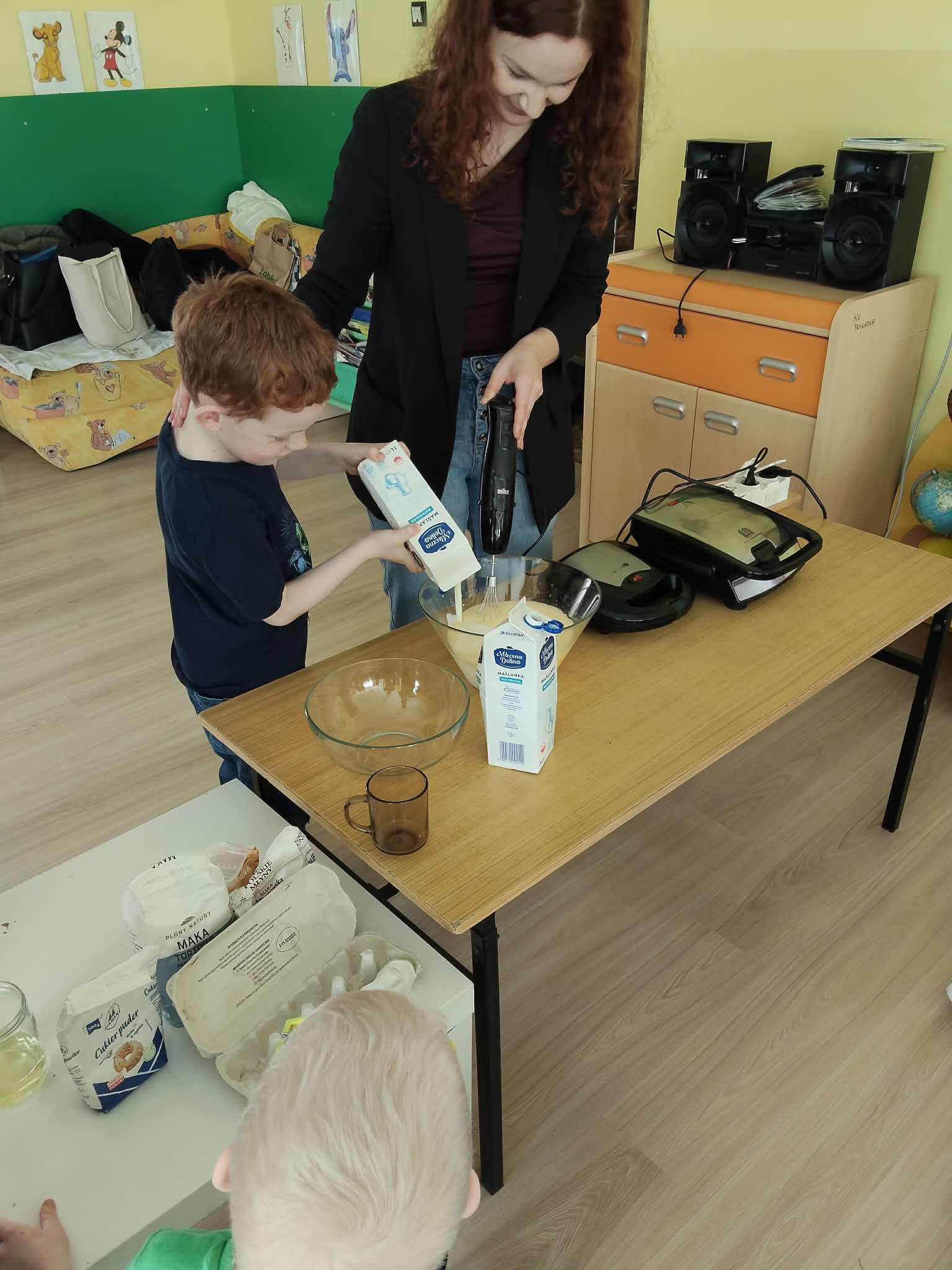 Osoba pomaga dziecku w przygotowaniu ciasta, podczas gdy dziecko wlewa składnik do miski stojącej na stole. Wokół znajdują się akcesoria kuchenne, a w tle widoczne są kolorowe elementy sali oraz sprzęt, co wskazuje na warsztaty kulinarne dla dzieci.