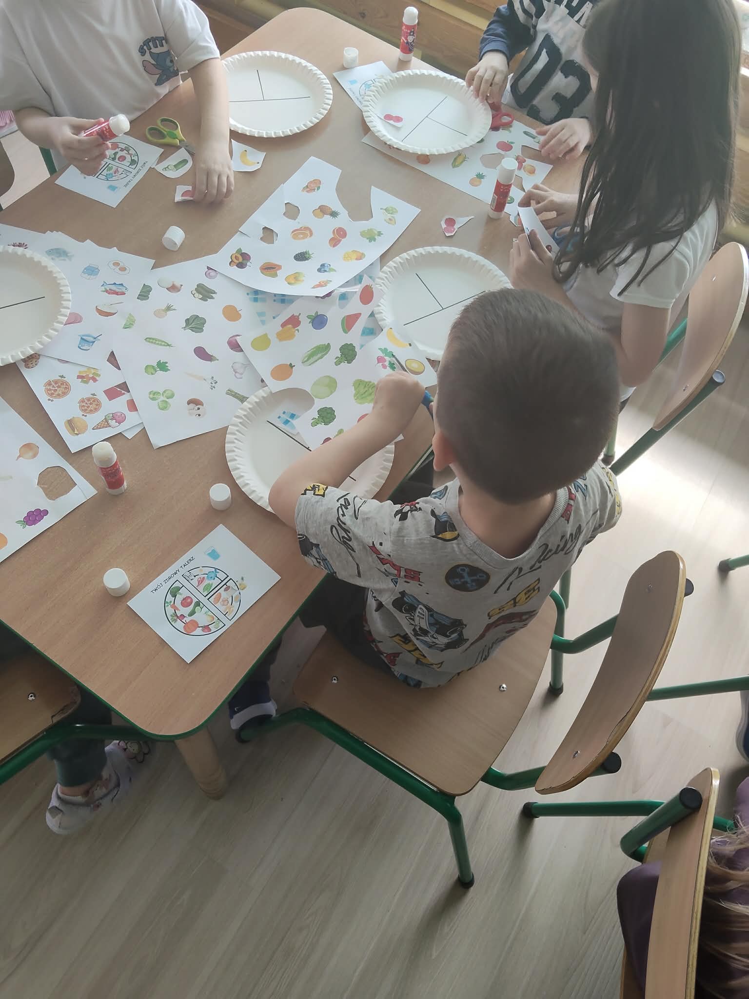 Dzieci siedzą przy stole i wykonują pracę plastyczną, tworząc talerze z podziałem na grupy produktów przy użyciu wycinanek z jedzeniem. Na stole znajdują się kartki, klej i nożyczki, a zajęcia mają charakter edukacyjny związany z nauką o żywieniu.