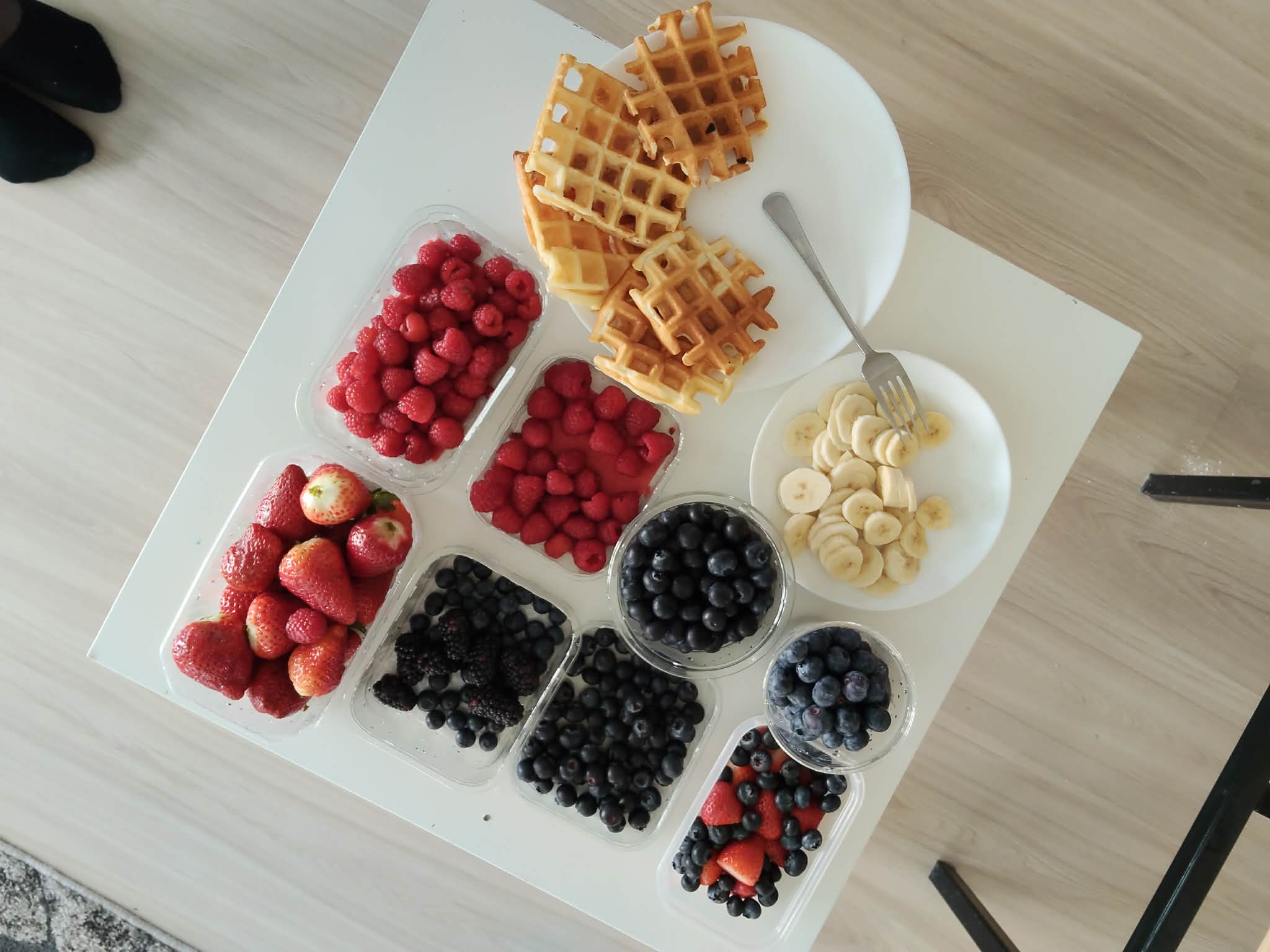 Stół z różnorodnymi produktami śniadaniowymi, na którym znajdują się gofry oraz talerz z pokrojonym bananem. Obok ustawione są pojemniki ze świeżymi owocami, takimi jak truskawki, maliny, borówki i jeżyny.