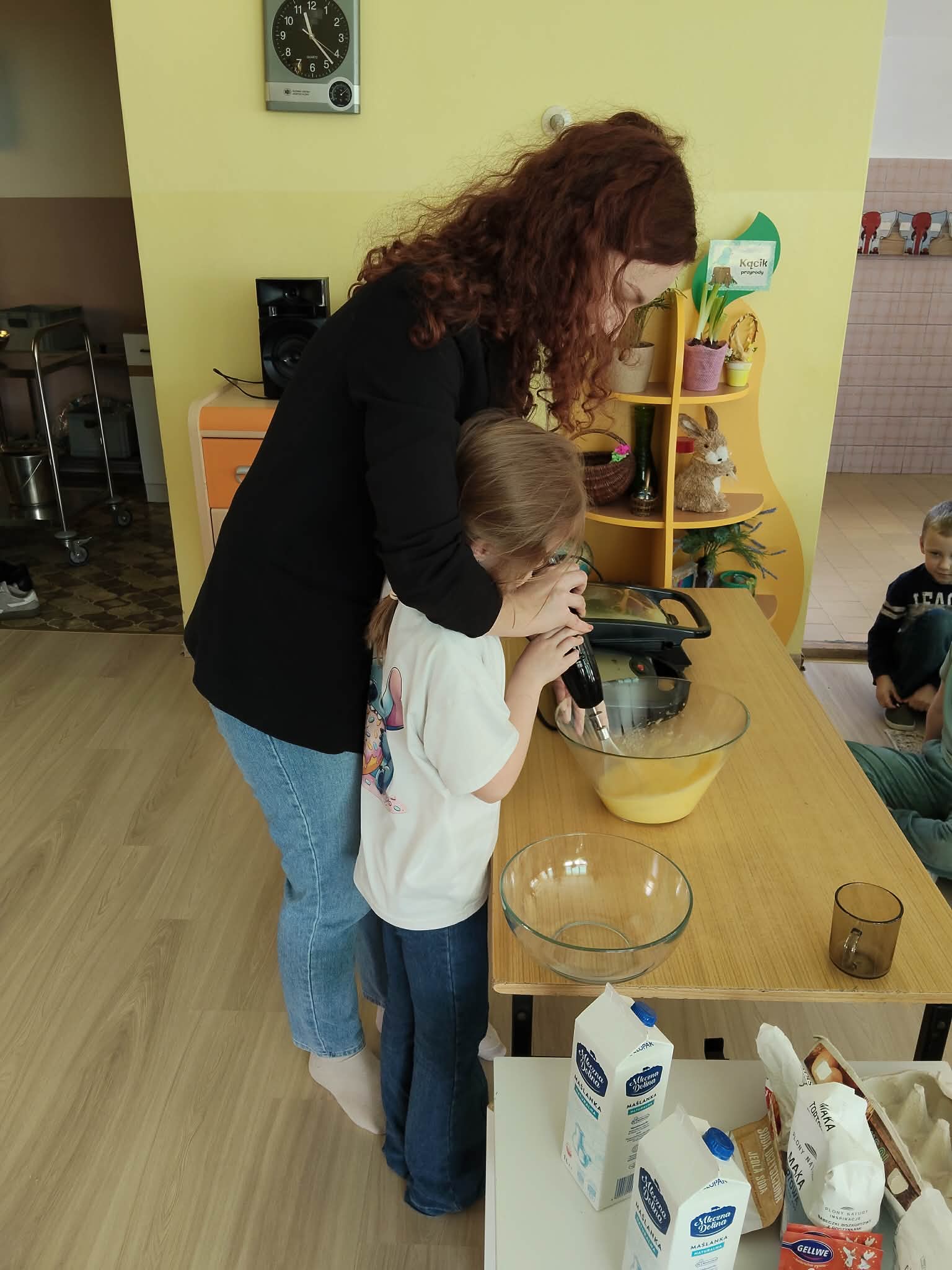 Osoba prowadzi zajęcia kulinarne, pomagając dziecku w użyciu miksera do przygotowania ciasta w misce na stole. Wokół znajdują się składniki i akcesoria do pieczenia, a w tle widoczne są elementy sali oraz inni uczestnicy zajęć.