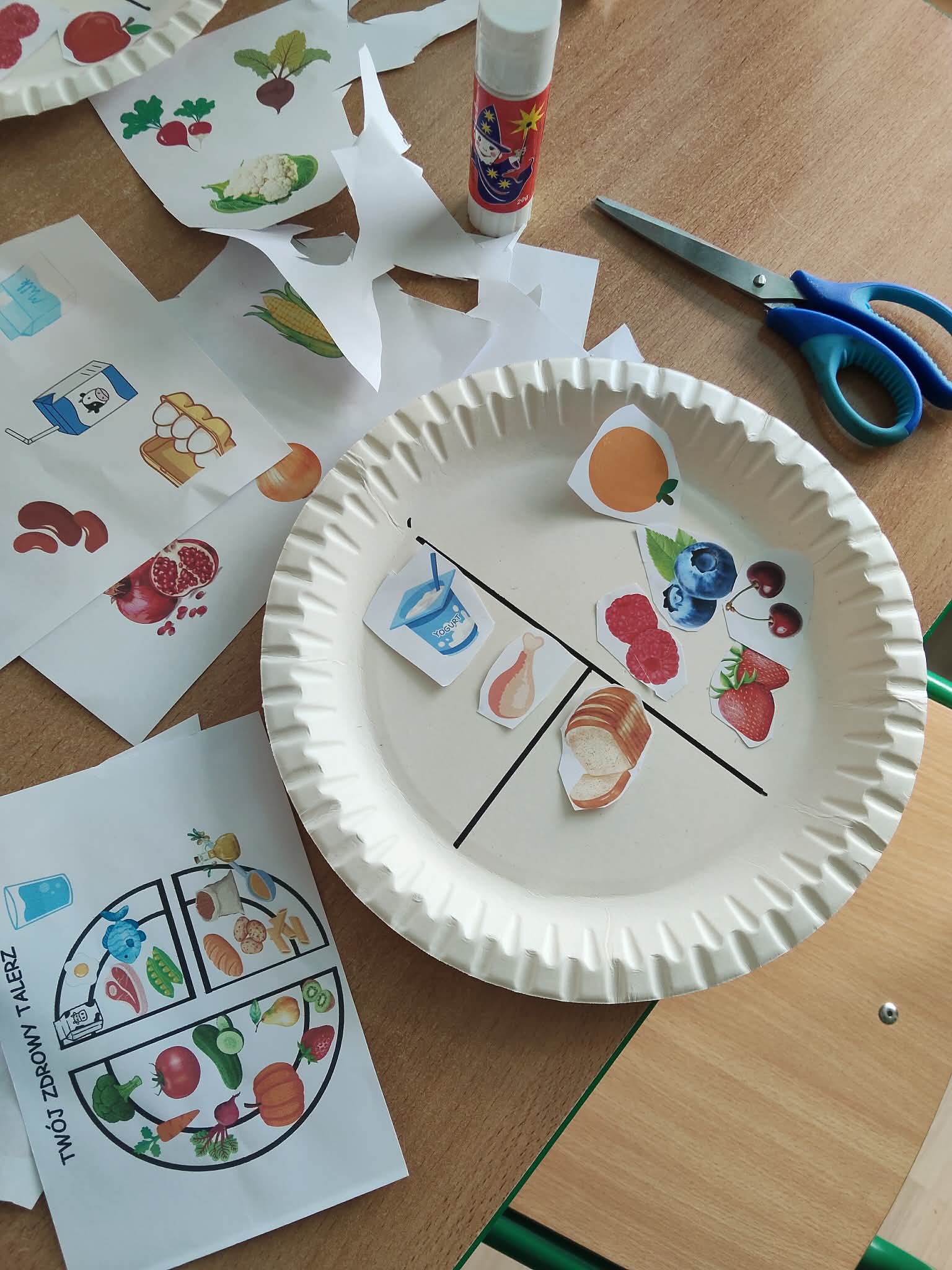 Praca plastyczna przedstawiająca talerz z podziałem na grupy produktów, wypełniony ilustracjami jedzenia. Obok znajdują się materiały do wykonania zadania, takie jak wycinanki, klej i nożyczki, co wskazuje na zajęcia edukacyjne dotyczące żywienia.