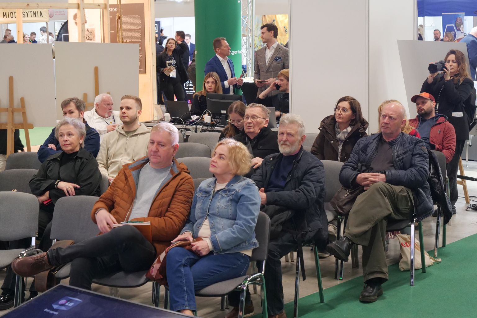 Uczestnicy siedzą na krzesłach i obserwują wydarzenie odbywające się poza kadrem. W tle widoczne są stanowiska z komputerami i sprzętem oraz osoby rozmawiające, co wskazuje na konferencję, prezentację lub wydarzenie targowe.
