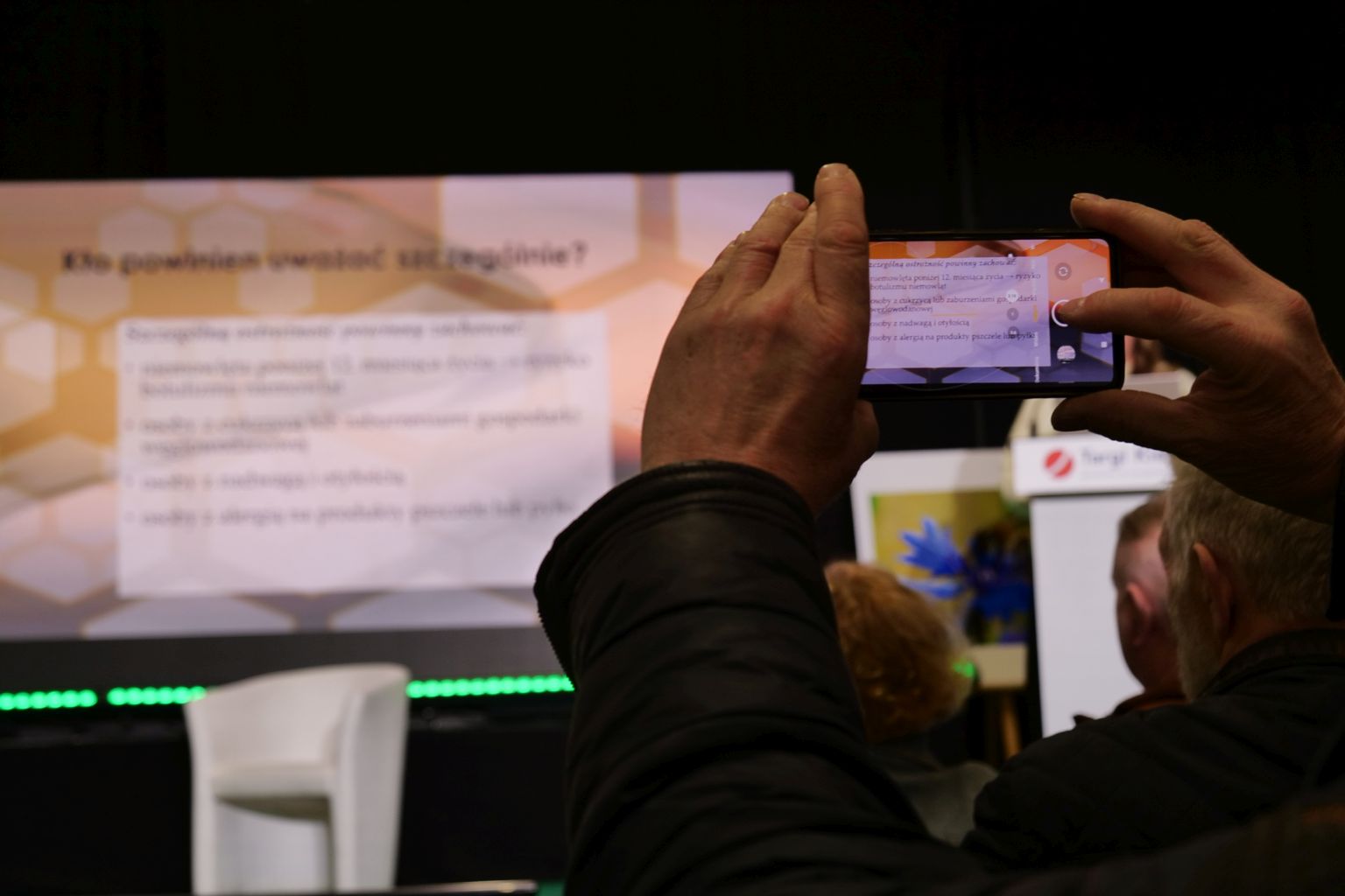 Uczestnicy znajdują się w sali konferencyjnej podczas prezentacji wyświetlanej na dużym ekranie. Jedna z osób rejestruje wydarzenie telefonem, a w tle widoczna jest scena z fotelem i mównicą. Całość ma charakter wydarzenia branżowego lub targowego.