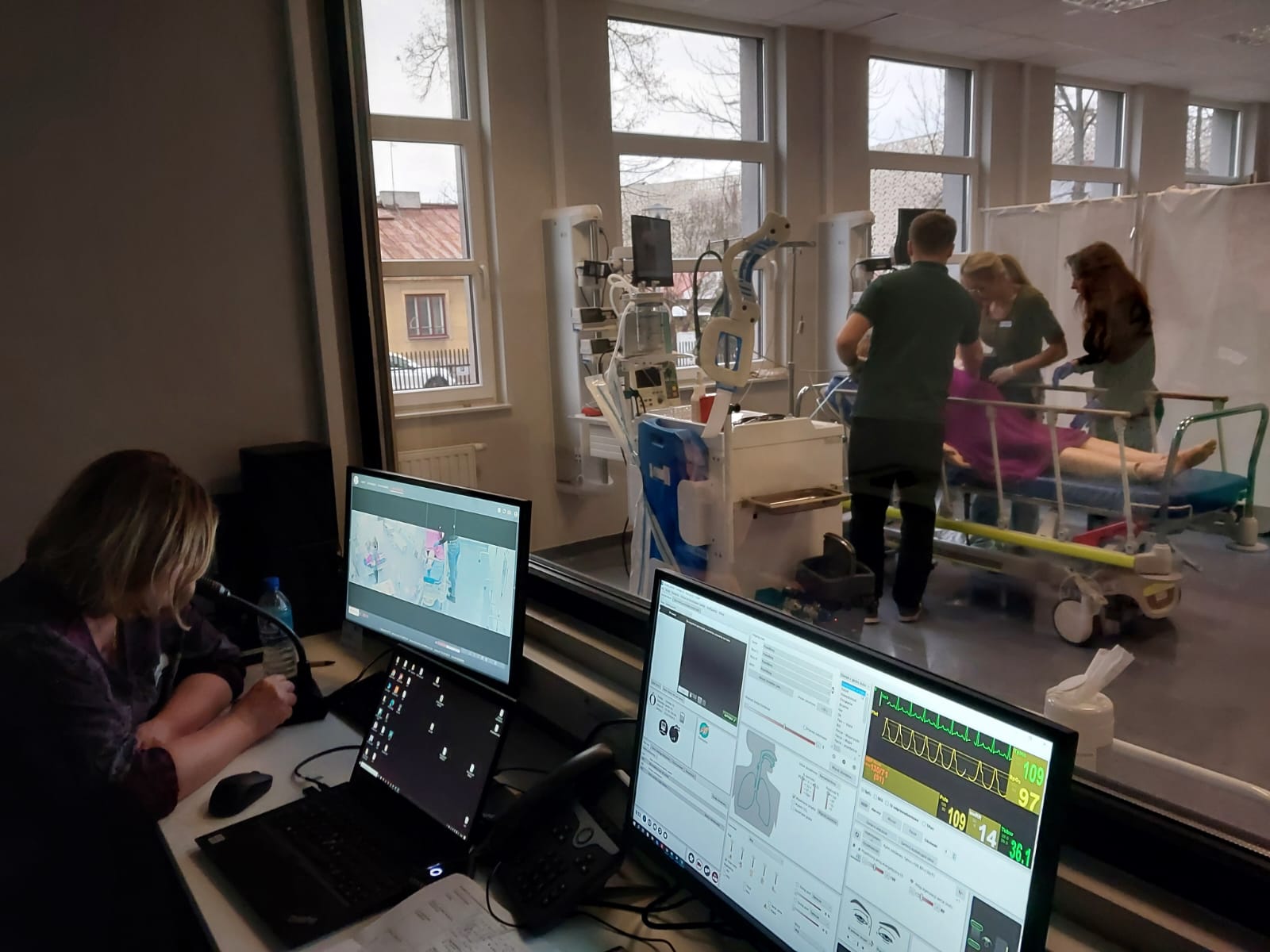 Widok na salę symulacji medycznej przez szybę, z pomieszczeniem sterowni na pierwszym planie. Osoba przy stanowisku komputerowym monitoruje przebieg ćwiczeń na ekranach, a w tle uczestnicy pracują przy łóżku z fantomem pacjenta. Sala wyposażona jest w sprzęt medyczny i urządzenia monitorujące, co wskazuje na realistyczne szkolenie.