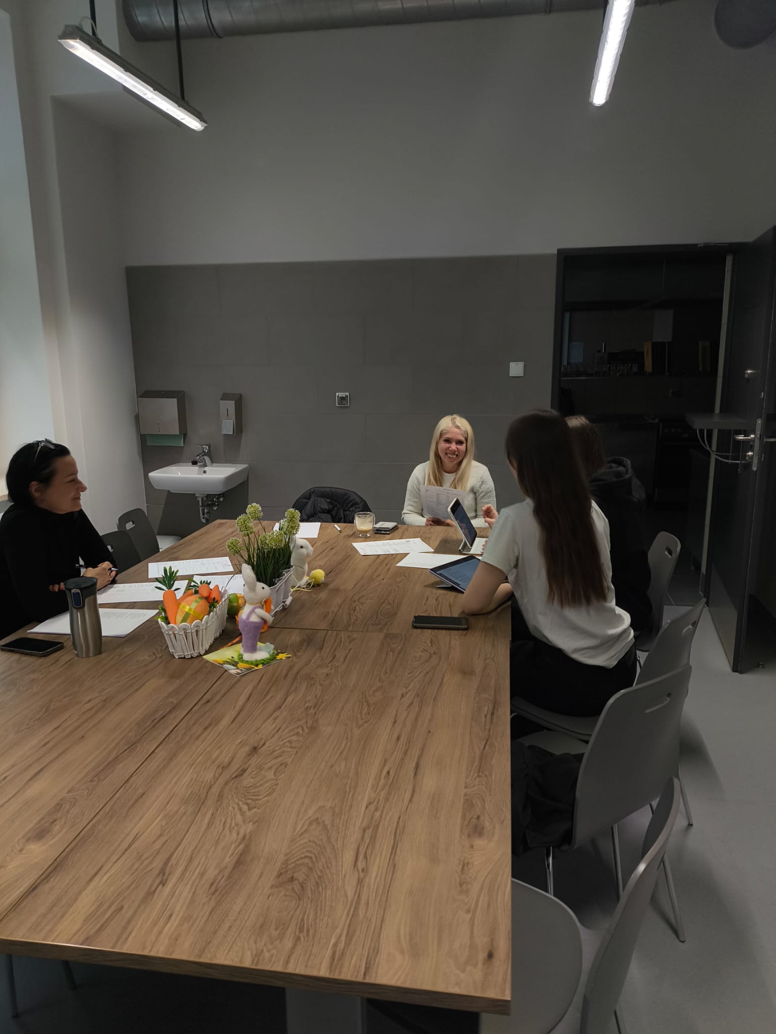 Cztery osoby siedzą przy długim stole w nowoczesnej sali spotkań i prowadzą rozmowę lub naradę. Na stole znajdują się dokumenty oraz kosz z owocami. Wnętrze ma industrialny charakter z szarymi ścianami, oświetleniem sufitowym i widocznymi instalacjami.