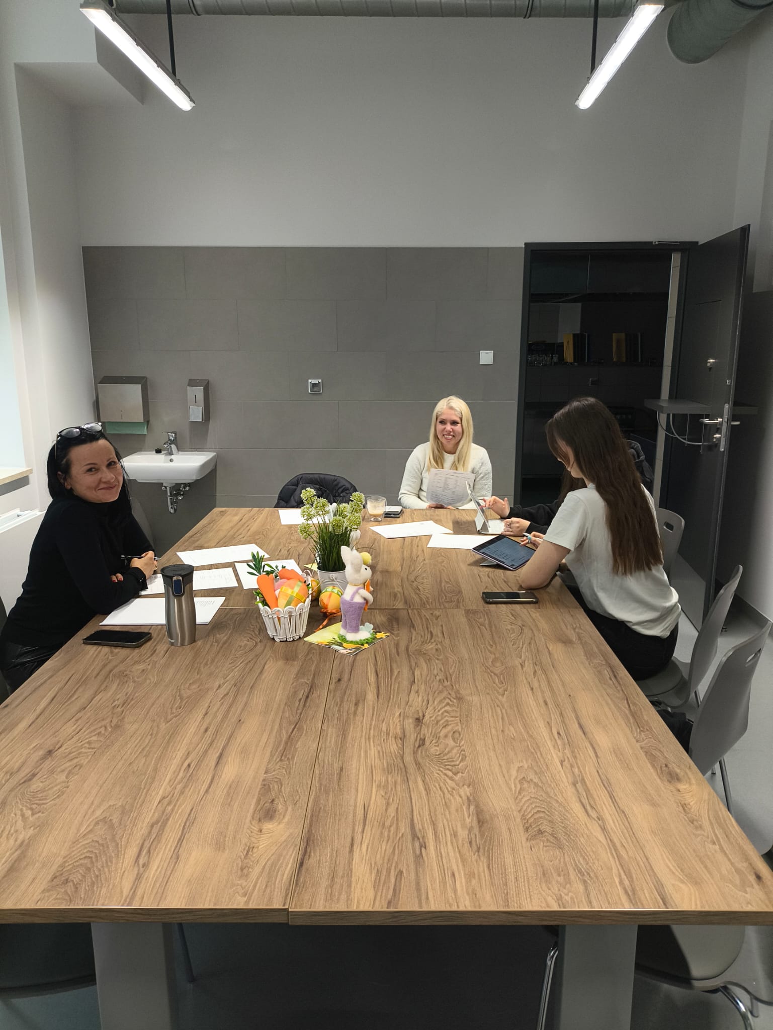 Cztery osoby siedzą przy długim stole w nowoczesnej sali spotkań i prowadzą rozmowę lub naradę. Na stole znajdują się dokumenty oraz kosz z owocami. Wnętrze ma industrialny charakter z szarymi ścianami, oświetleniem sufitowym i widocznymi instalacjami.