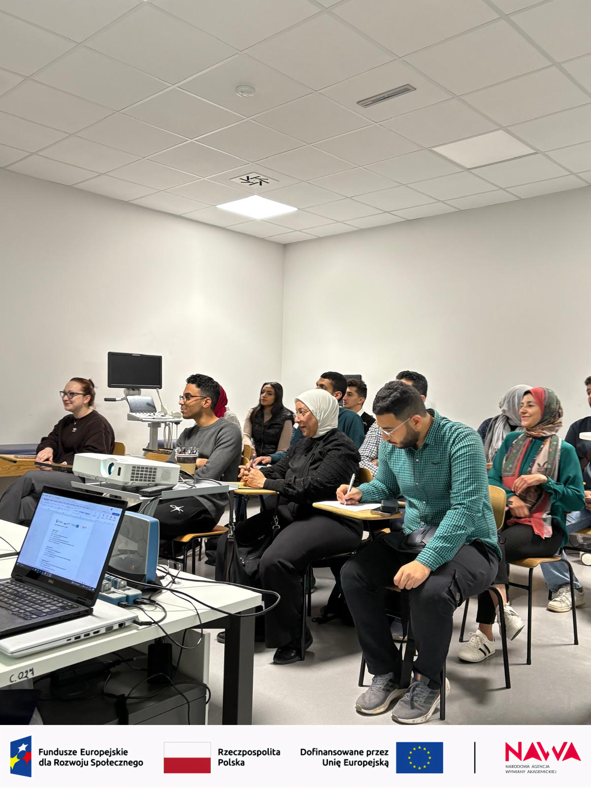 Sala dydaktyczna z uczestnikami siedzącymi w rzędach i obserwującymi zajęcia prowadzone z przodu. Na pierwszym planie widoczne jest stanowisko z laptopem i projektorem wykorzystywanym do prezentacji. Pomieszczenie ma jasne ściany, oświetlenie sufitowe oraz elementy wyposażenia typowe dla zajęć edukacyjnych.