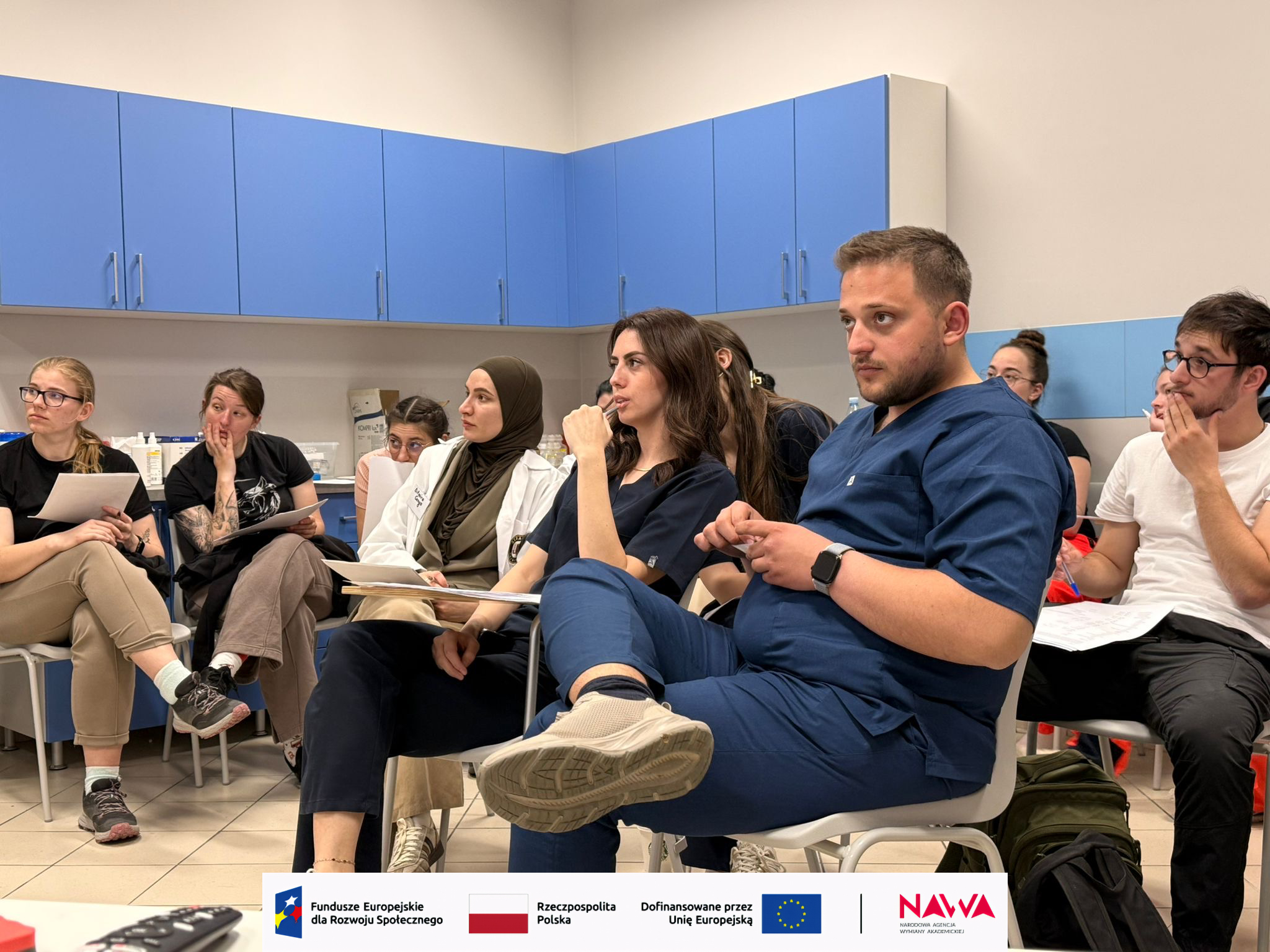 Uczestnicy siedzą w półkolu w sali dydaktycznej i biorą udział w spotkaniu lub dyskusji. W tle widoczne są szafki i blat z wyposażeniem, co sugeruje przestrzeń edukacyjną lub laboratoryjną. Część osób trzyma materiały, a całość ma charakter zajęć lub warsztatów.