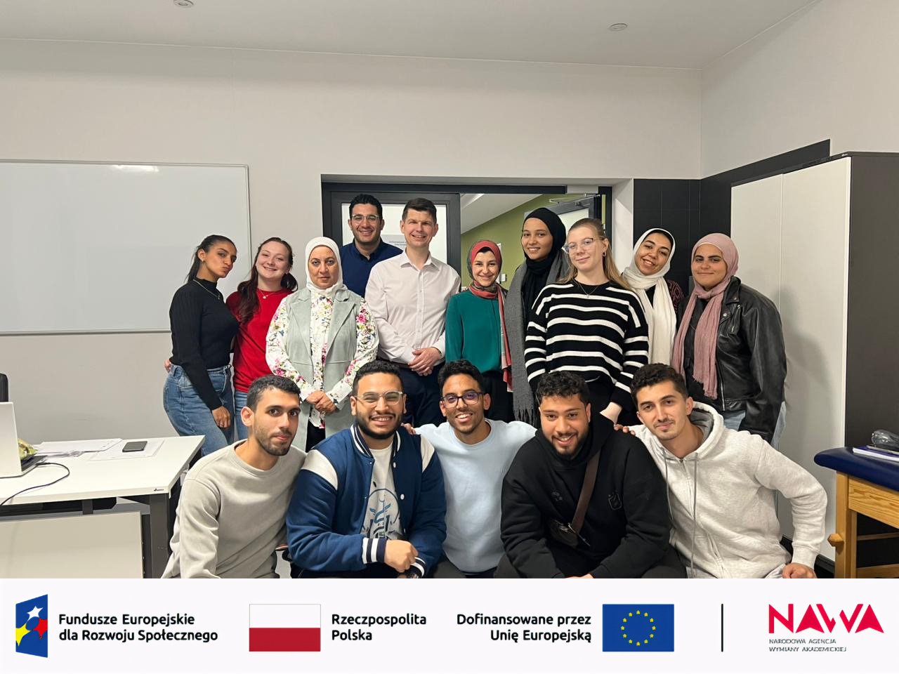 Grupa uczestników znajduje się w sali dydaktycznej i pozuje do wspólnego zdjęcia, część osób stoi, a część siedzi z przodu. W tle widoczna jest tablica i drzwi, a całość ma charakter zajęć lub projektu edukacyjnego. Na dole znajdują się logotypy związane z finansowaniem i organizacją przedsięwzięcia.