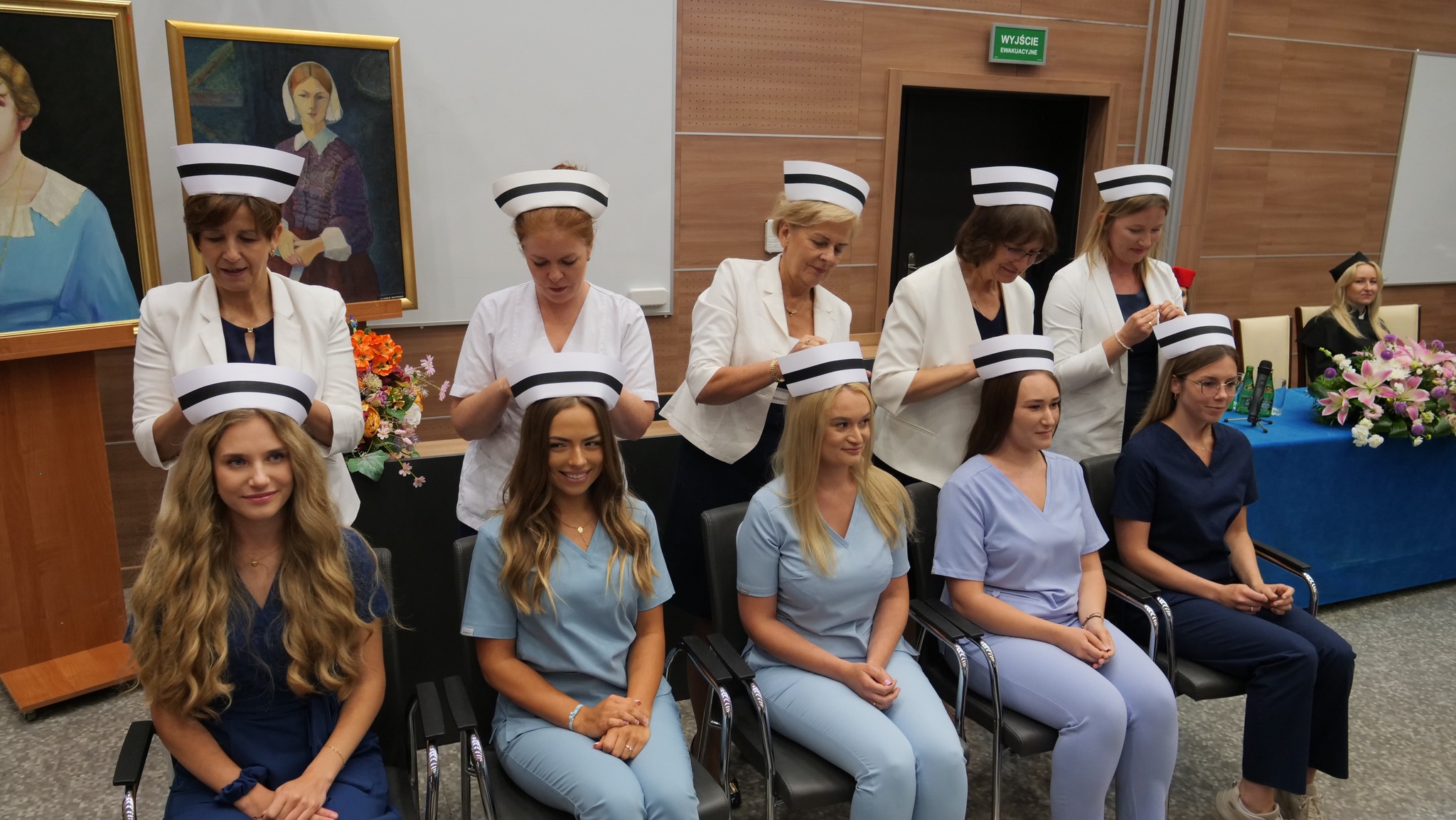 studentki siedzą na krzesłach w strojach pielęgniarek. Wykładowcy zakładają studentkom czepki pielęgniarskie. Wykładowcy Agnieszka Kaleta, Małgorzata Dudek, Ewa Kozłowska, Małgorzata Kaczmarczyk, Marta Kordyzon. Przy stole siedzi Prodziekan Dorota Rębak