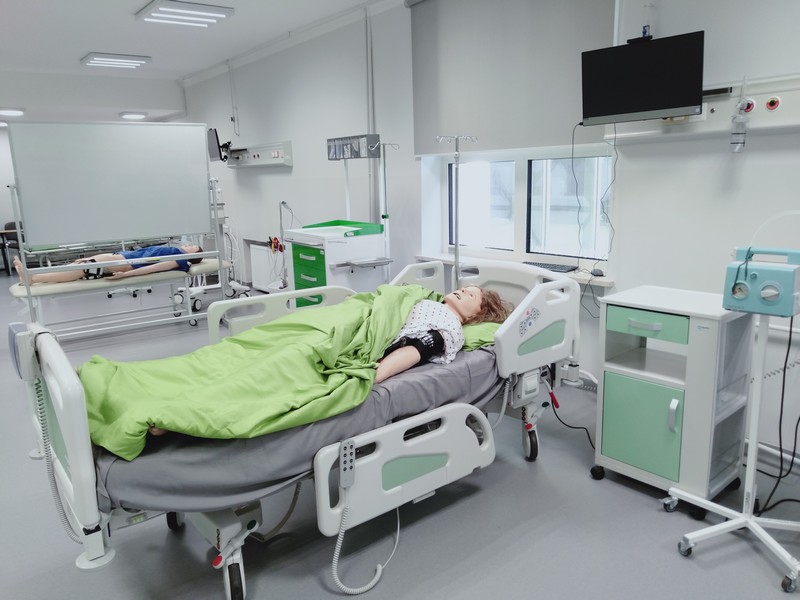 Osoba leżąca na łóżku szpitalnym w nowoczesnej sali medycznej, przykryta zieloną pościelą; wokół znajdują się aparatura medyczna, szafki, monitor na ścianie oraz inne łóżka szpitalne