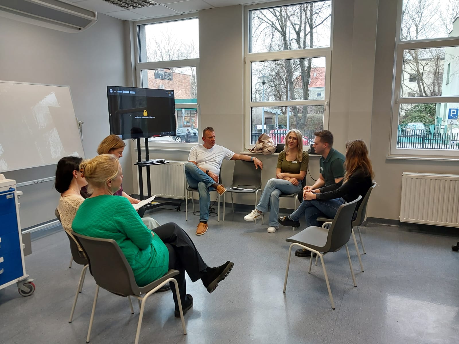 Grupa uczestników siedzi w kręgu w jasnej sali i prowadzi rozmowę lub zajęcia warsztatowe. Wnętrze ma nowoczesny charakter z dużymi oknami, tablicą oraz ekranem, a naturalne światło podkreśla otwartą i sprzyjającą dyskusji atmosferę.