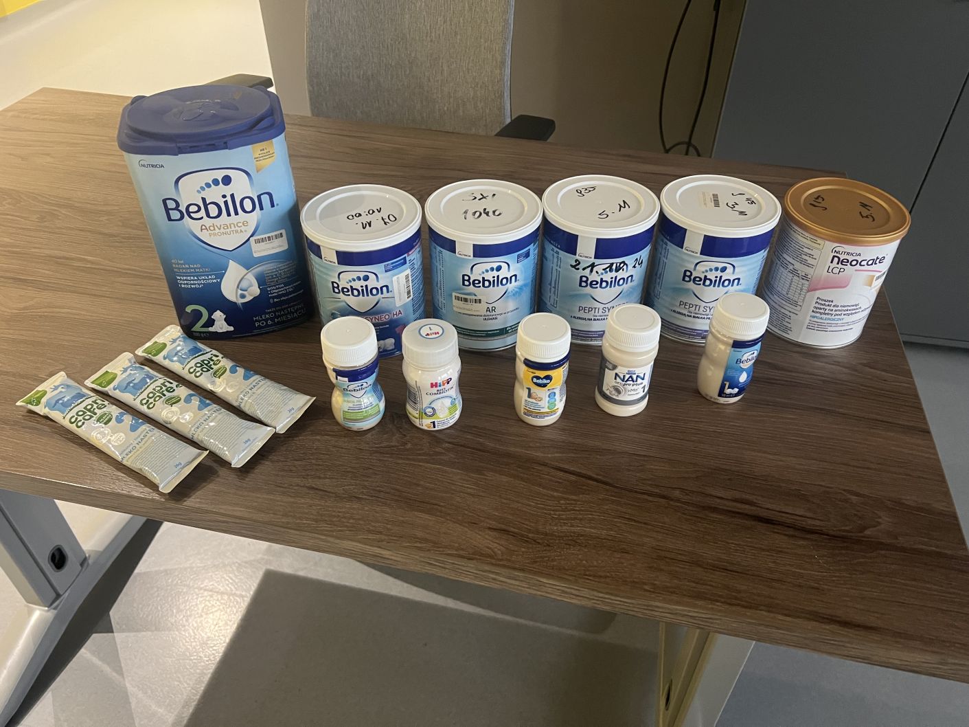 Na drewnianym stole ustawiono różne preparaty i produkty żywieniowe dla niemowląt, w tym puszki mleka modyfikowanego, saszetki oraz małe butelki z gotowymi produktami. Materiały zostały przygotowane do prezentacji lub zajęć edukacyjnych związanych z żywieniem dzieci.