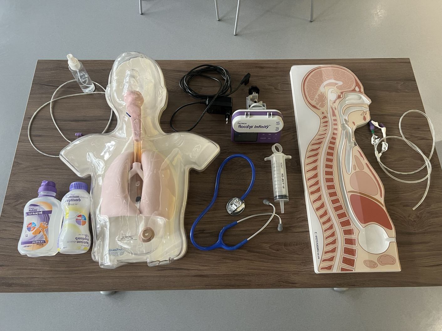 Zestaw materiałów i sprzętu wykorzystywanego podczas szkolenia medycznego, ułożony na stole w sali dydaktycznej. Widoczne są modele anatomiczne, stetoskop, strzykawka, pompa do żywienia oraz przewody i akcesoria medyczne służące do ćwiczeń praktycznych.