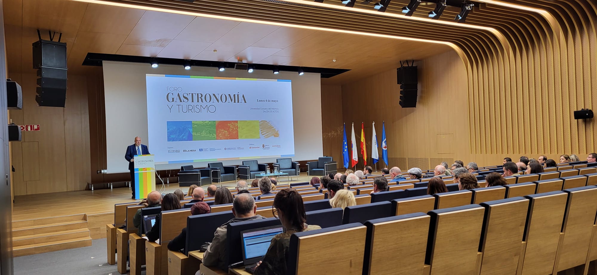 Sala konferencyjna z drewnianym wykończeniem i rzędami stopniowanych siedzeń, w której uczestnicy słuchają prezentacji prowadzonej z mównicy. Na dużym ekranie wyświetlany jest slajd wydarzenia „Foro Gastronomía y Turismo”, a obok sceny widoczne są flagi oraz elementy identyfikacji wydarzenia. Wnętrze ma nowoczesny, profesjonalny charakter.