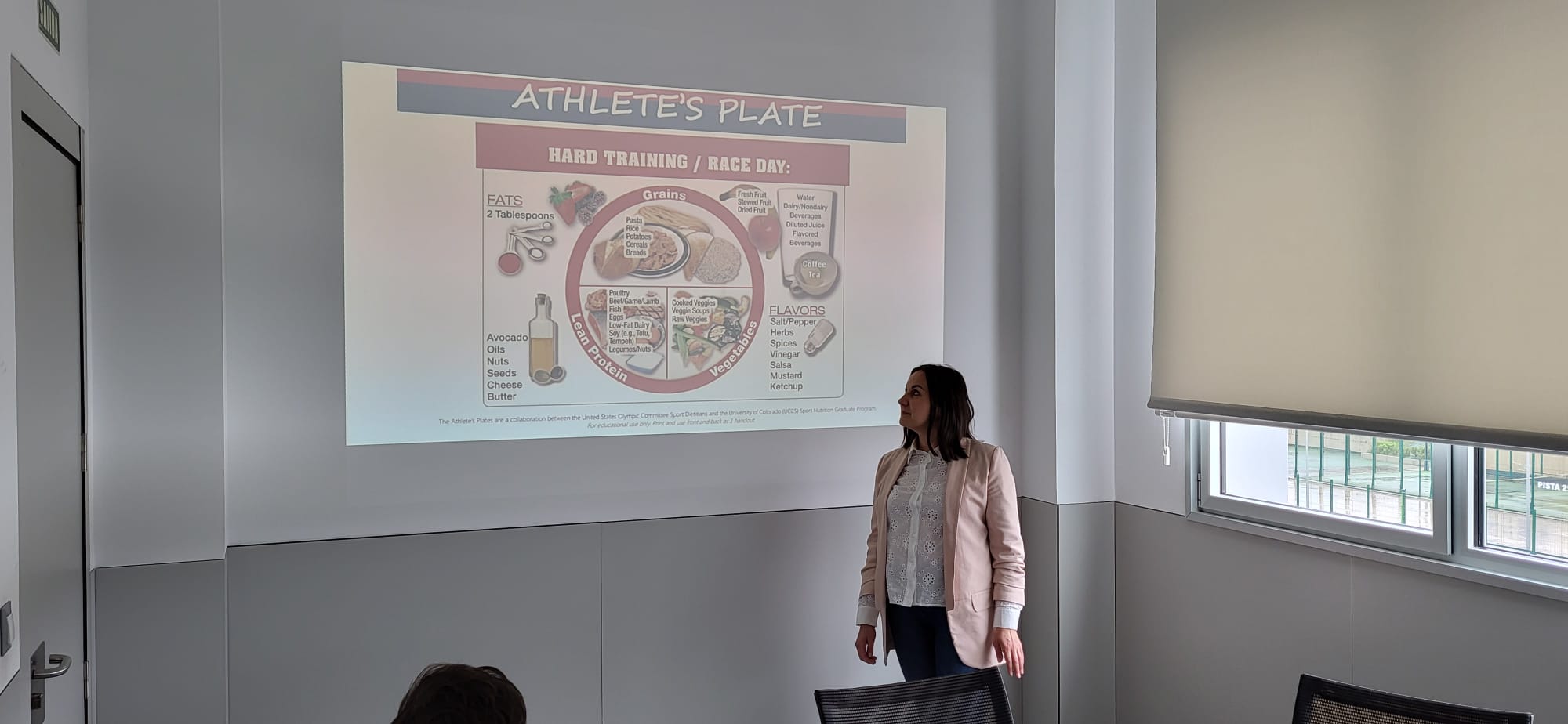 Osoba prowadzi prezentację przy slajdzie dotyczącym żywienia sportowego i schematu „Athlete’s Plate”. Na ekranie widoczny jest podział produktów spożywczych na kategorie związane z dietą podczas intensywnego treningu. Sala ma jasne ściany i okno wpuszczające światło dzienne, co nadaje wnętrzu nowoczesny charakter.