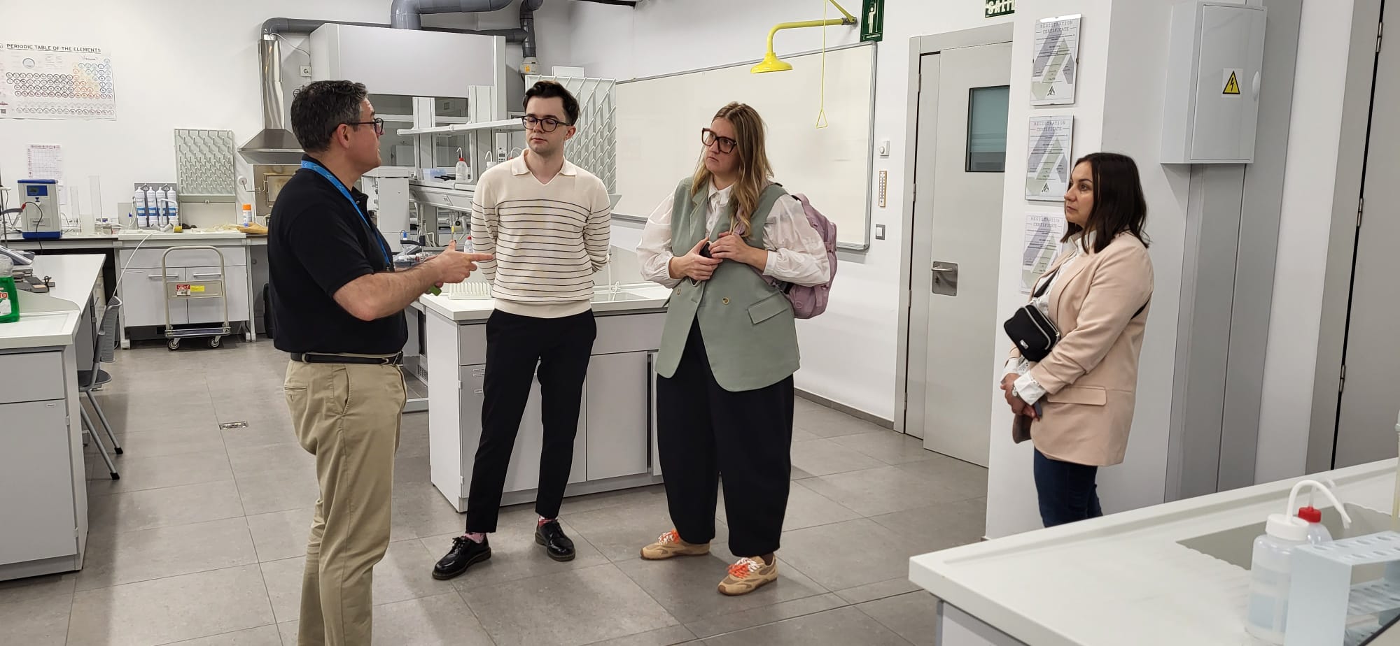 Uczestnicy rozmawiają w laboratorium lub sali dydaktycznej wyposażonej w stanowiska i materiały laboratoryjne. Jedna z osób prowadzi rozmowę lub prezentację, gestykulując podczas wyjaśnień. W tle widoczne są elementy wyposażenia pracowni, tablica układu okresowego oraz oznaczenia bezpieczeństwa.