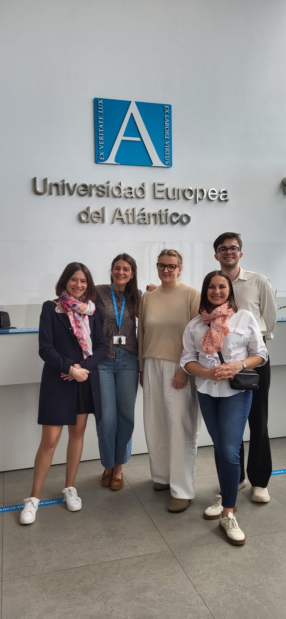 Grupa uczestników stoi przed ścianą z logotypem i nazwą Universidad Europea del Atlántico, pozując do wspólnego zdjęcia. Wnętrze jest jasne i nowoczesne, co wskazuje na akademicki lub konferencyjny charakter miejsca.