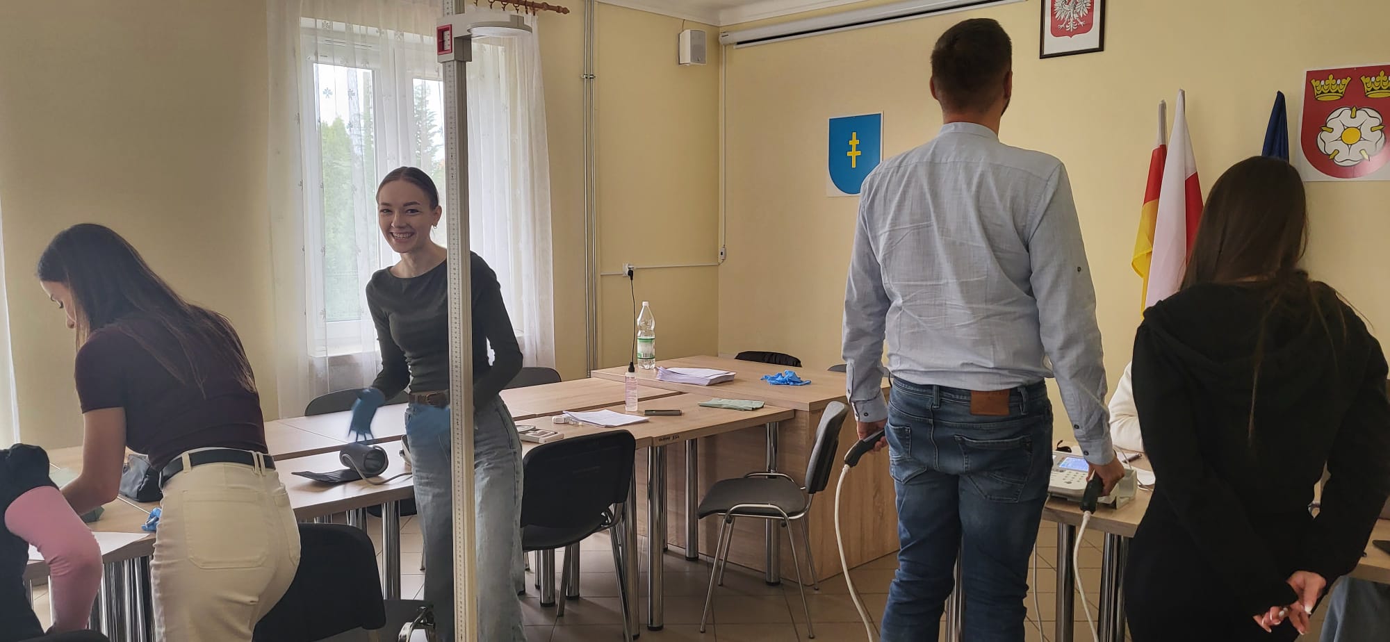 Uczestnicy znajdują się w sali o charakterze edukacyjnym lub organizacyjnym, wykonując różne zadania przy ustawionych stołach. Na blatach widoczne są dokumenty i akcesoria, a w tle znajdują się flagi oraz emblematy na ścianie. Pomieszczenie jest jasne, z dostępem do naturalnego światła z okna.