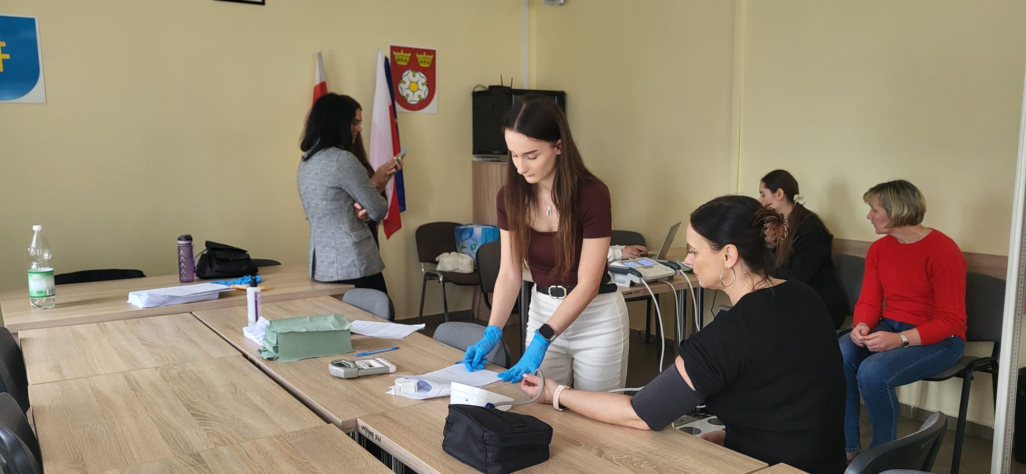 W sali o charakterze edukacyjnym lub zdrowotnym wykonywany jest pomiar ciśnienia tętniczego przy stole z dokumentami i sprzętem medycznym. Jedna z osób przeprowadza badanie, a w tle inni uczestnicy korzystają z laptopa lub oczekują na swoją kolej. Pomieszczenie ma jasne ściany oraz elementy dekoracyjne, takie jak flagi i herb.